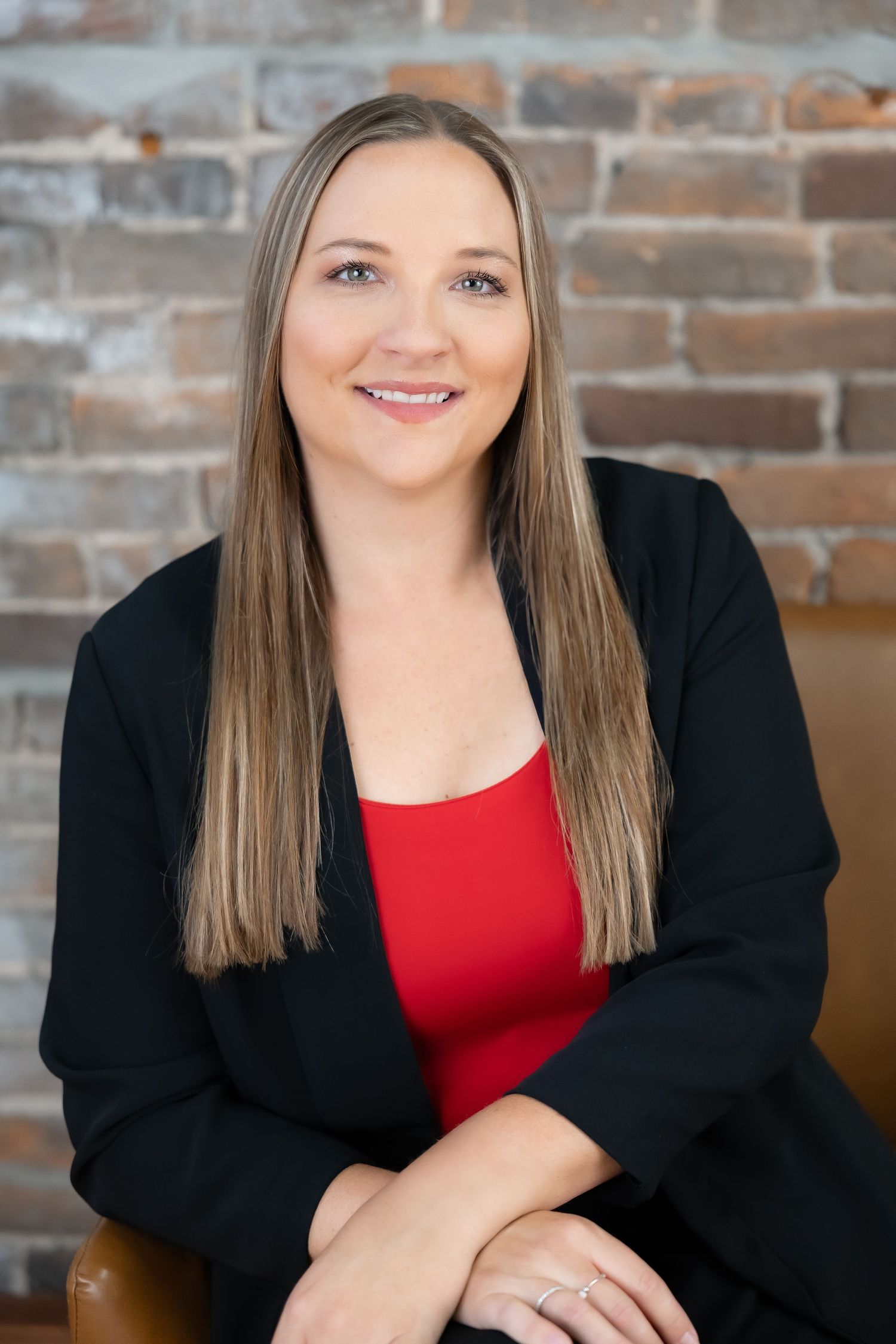 Business portrait featuring black blazer and red top against textured brick background.