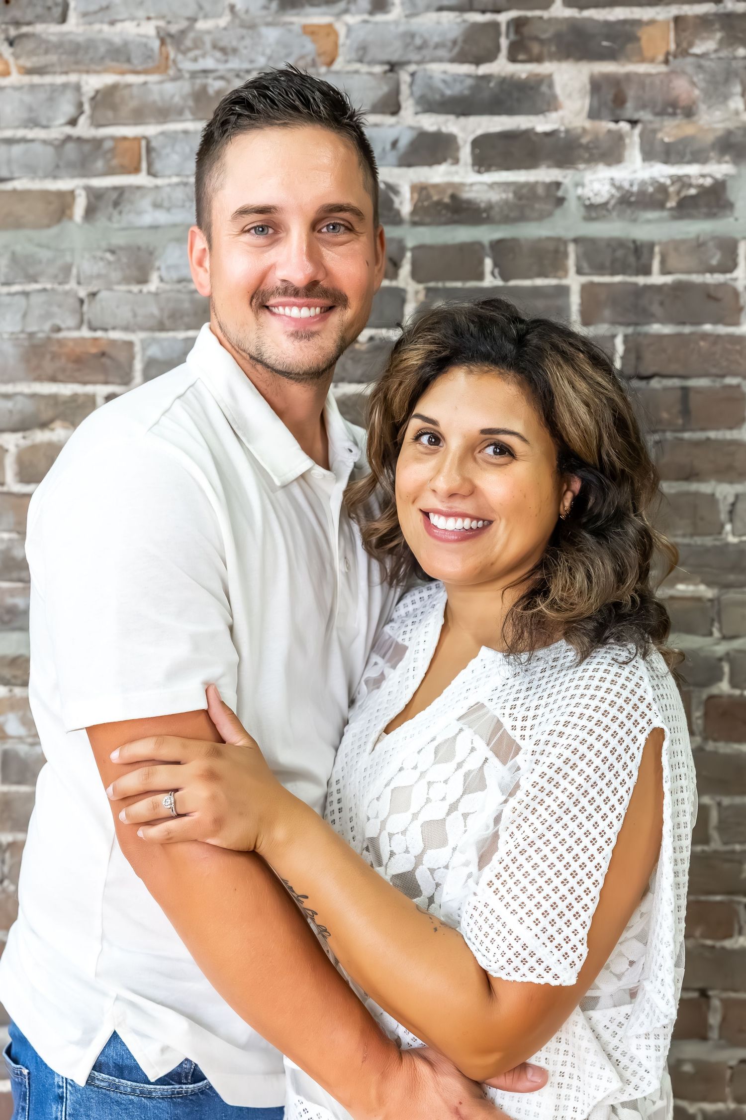 A couple dressed in white and blue embraces while smiling against an exposed brick wall.