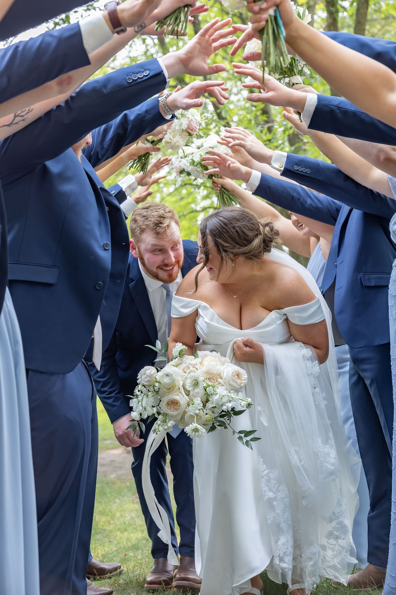 Wedding party in navy blue suits forms a tunnel with raised arms as newlyweds pass underneath with white bouquet.