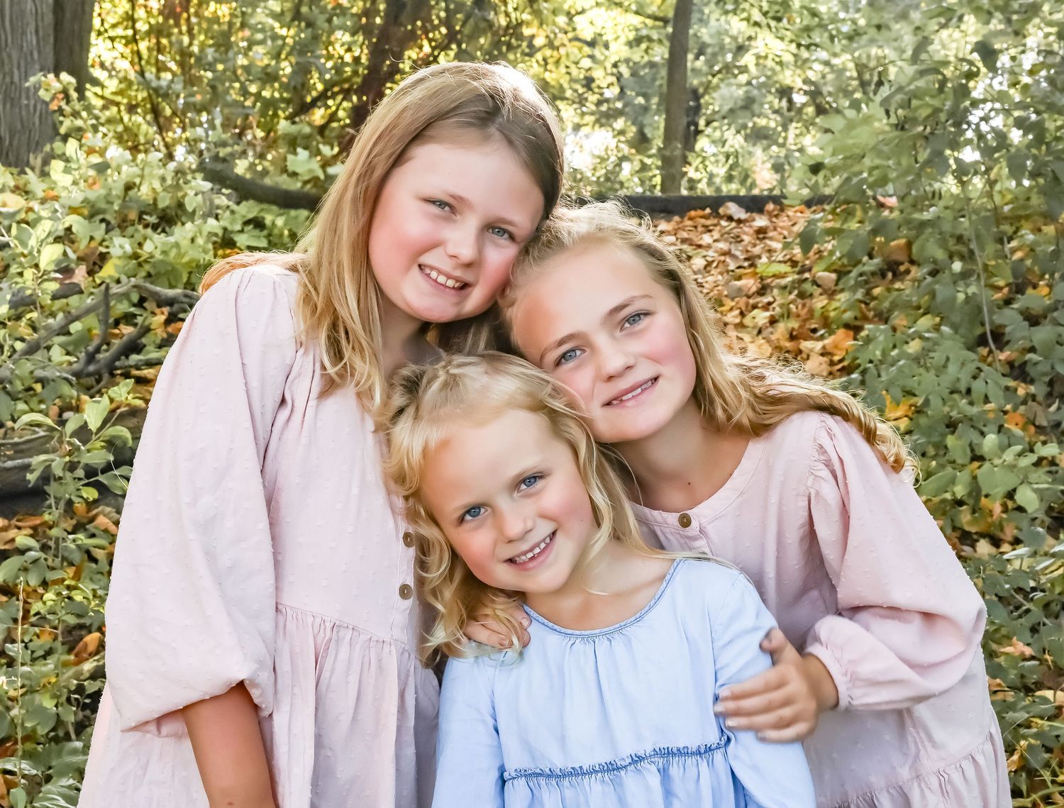 Three sisters in matching pink dresses pose together outdoors in an autumn setting.