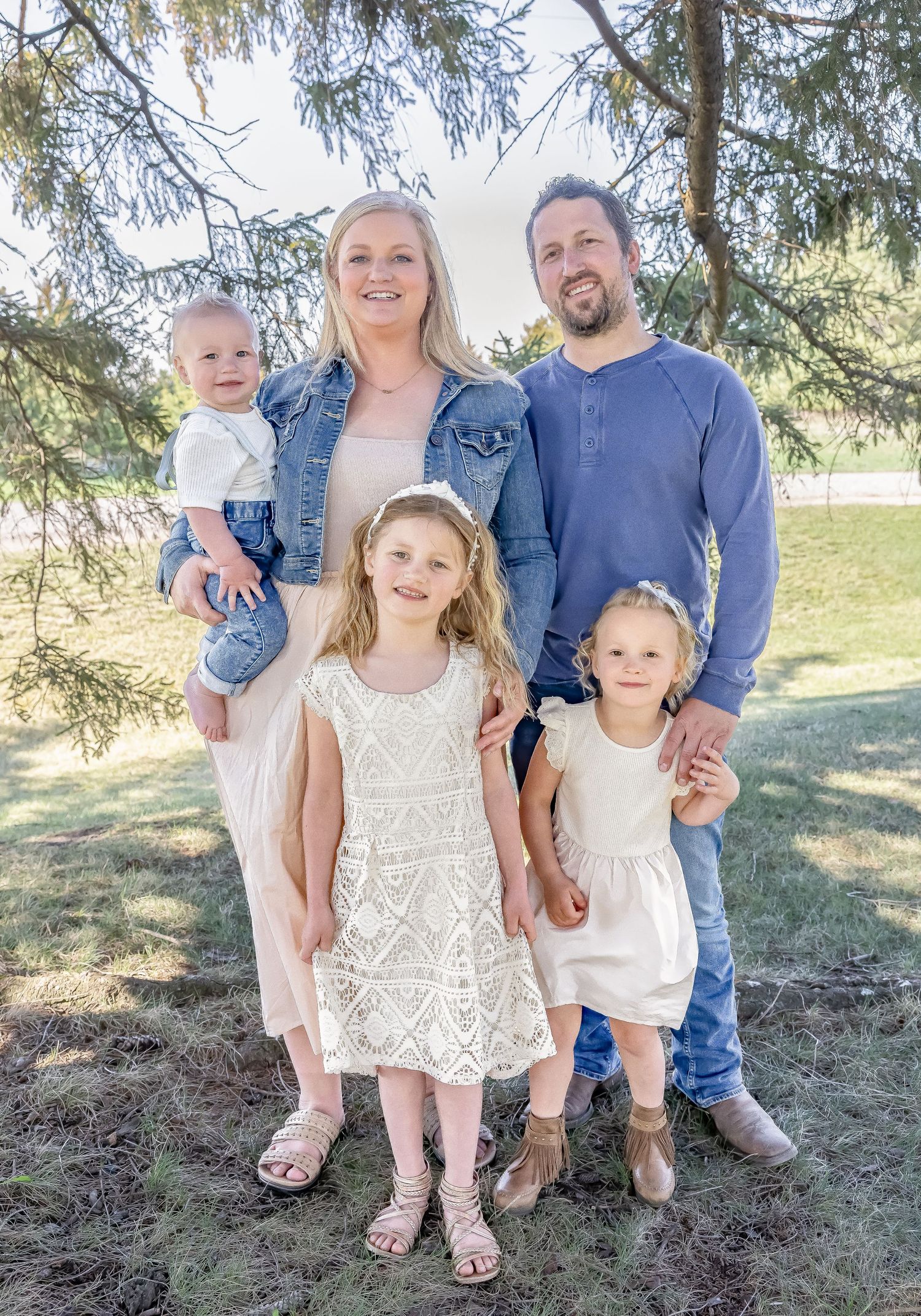 A family in coordinating white and blue outfits poses together outdoors under evergreen trees.
