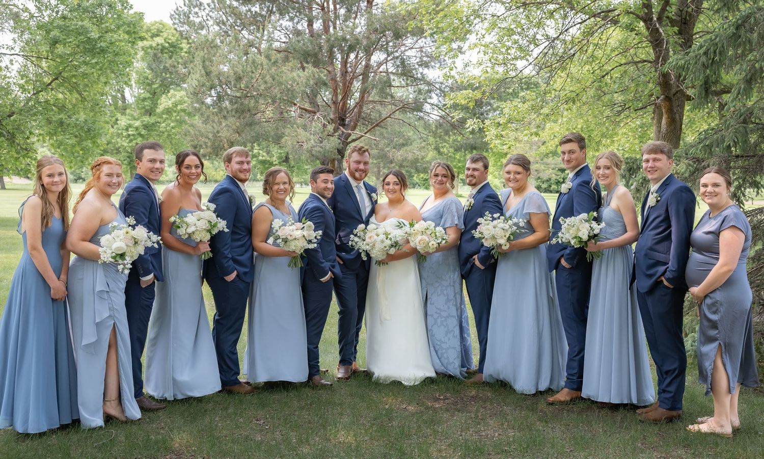 Wedding party in light blue bridesmaid dresses and navy suits pose together on grassy lawn under trees.