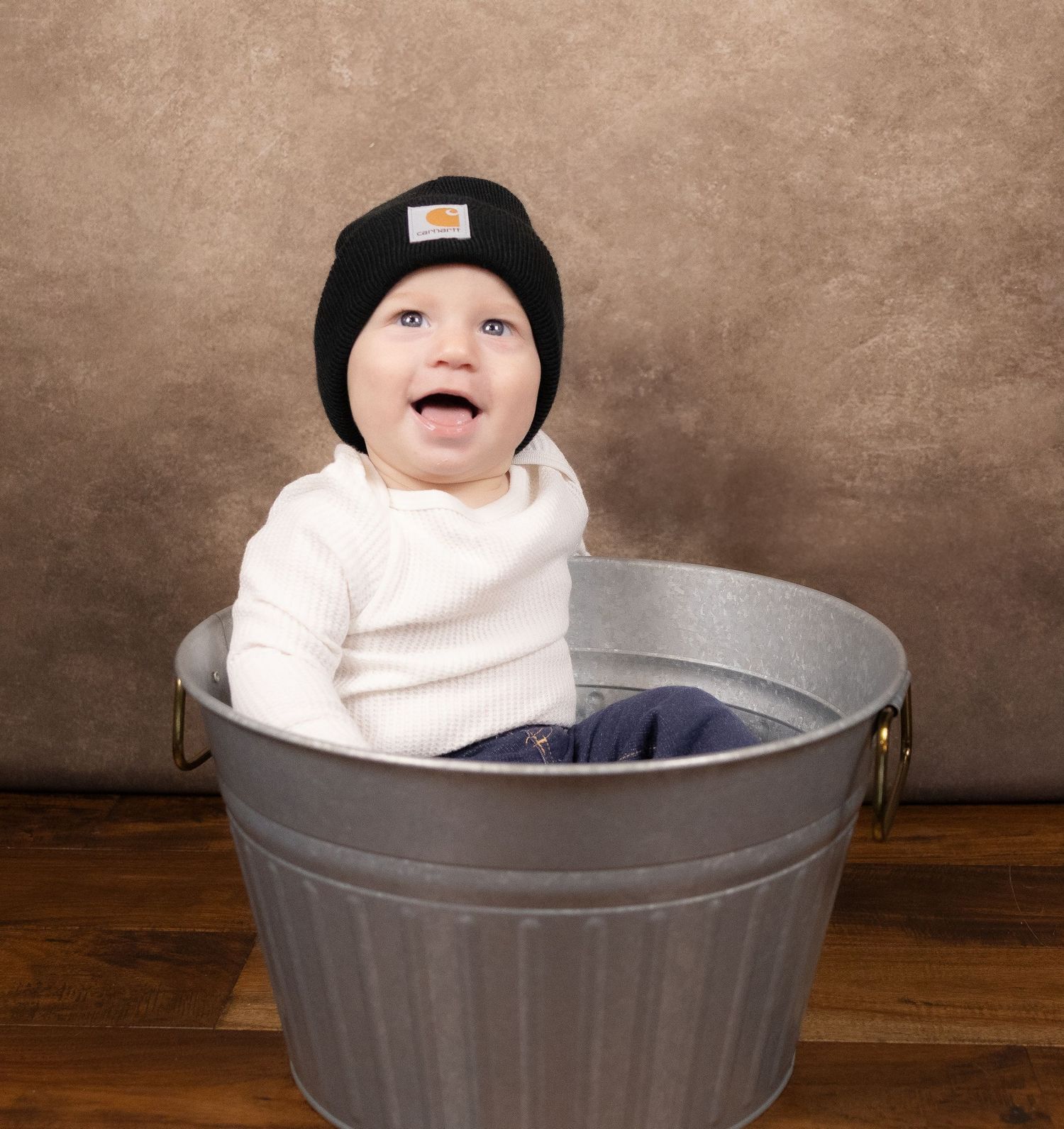 Baby wearing black beanie and white shirt sits happily in metal bucket during photo session.