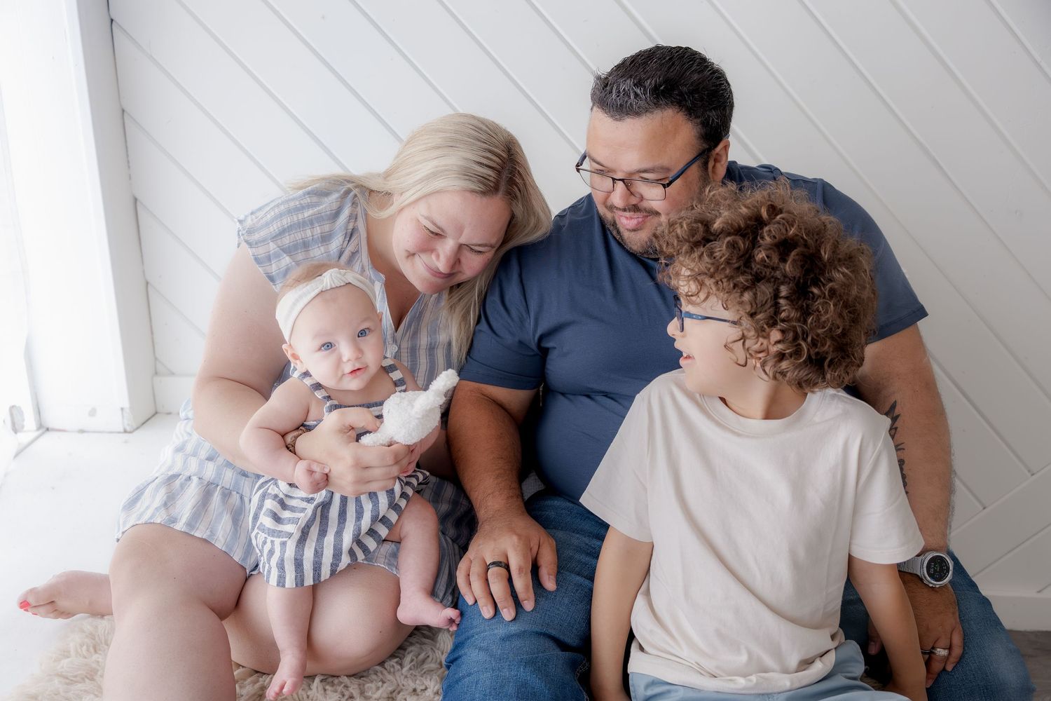 Happy family of four sitting together, parents smiling at baby and curly-haired toddler in bright white room.
