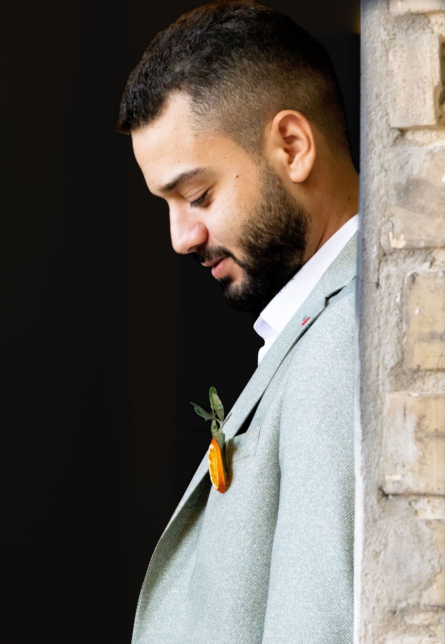 A groom in a light gray suit smiles while wearing a yellow boutonniere against a dark background.