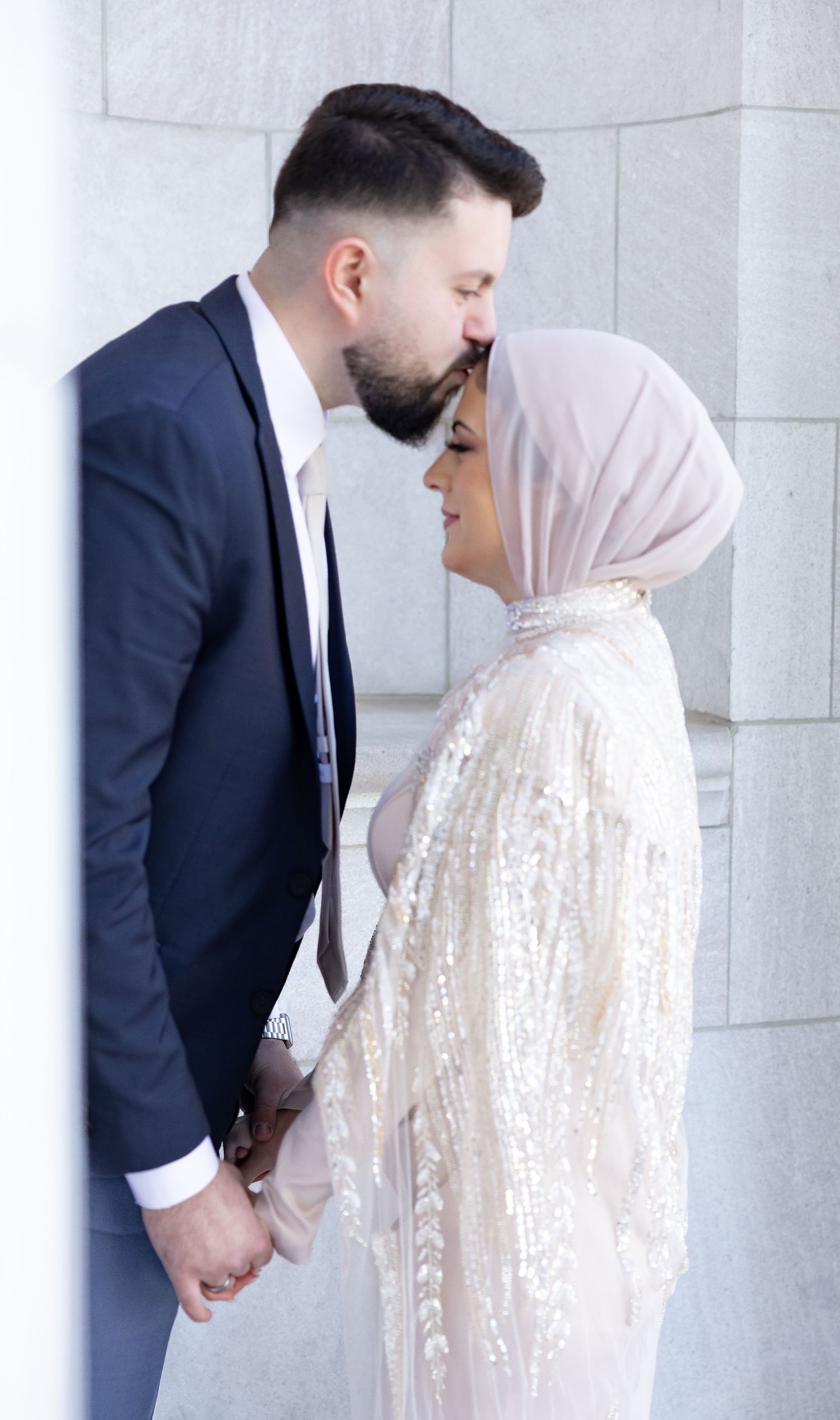 A couple in wedding attire share an intimate moment against a white stone wall.