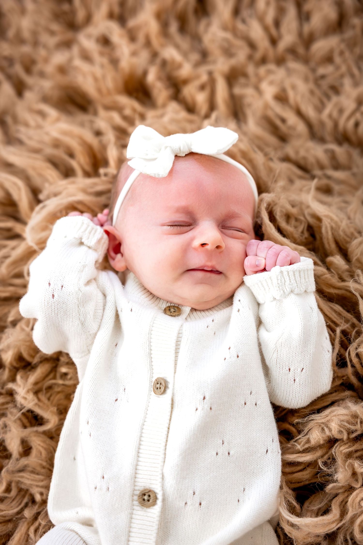 Sleeping baby wearing white knit cardigan with bow headband rests on fluffy brown blanket.