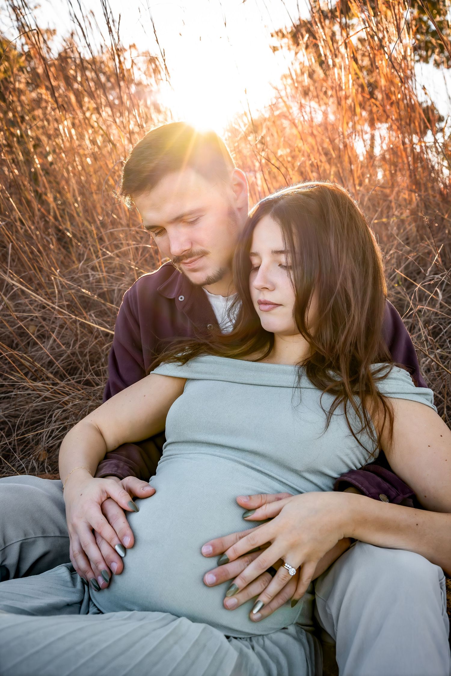 Romantic maternity portrait with sunburst through tall grass at golden hour.