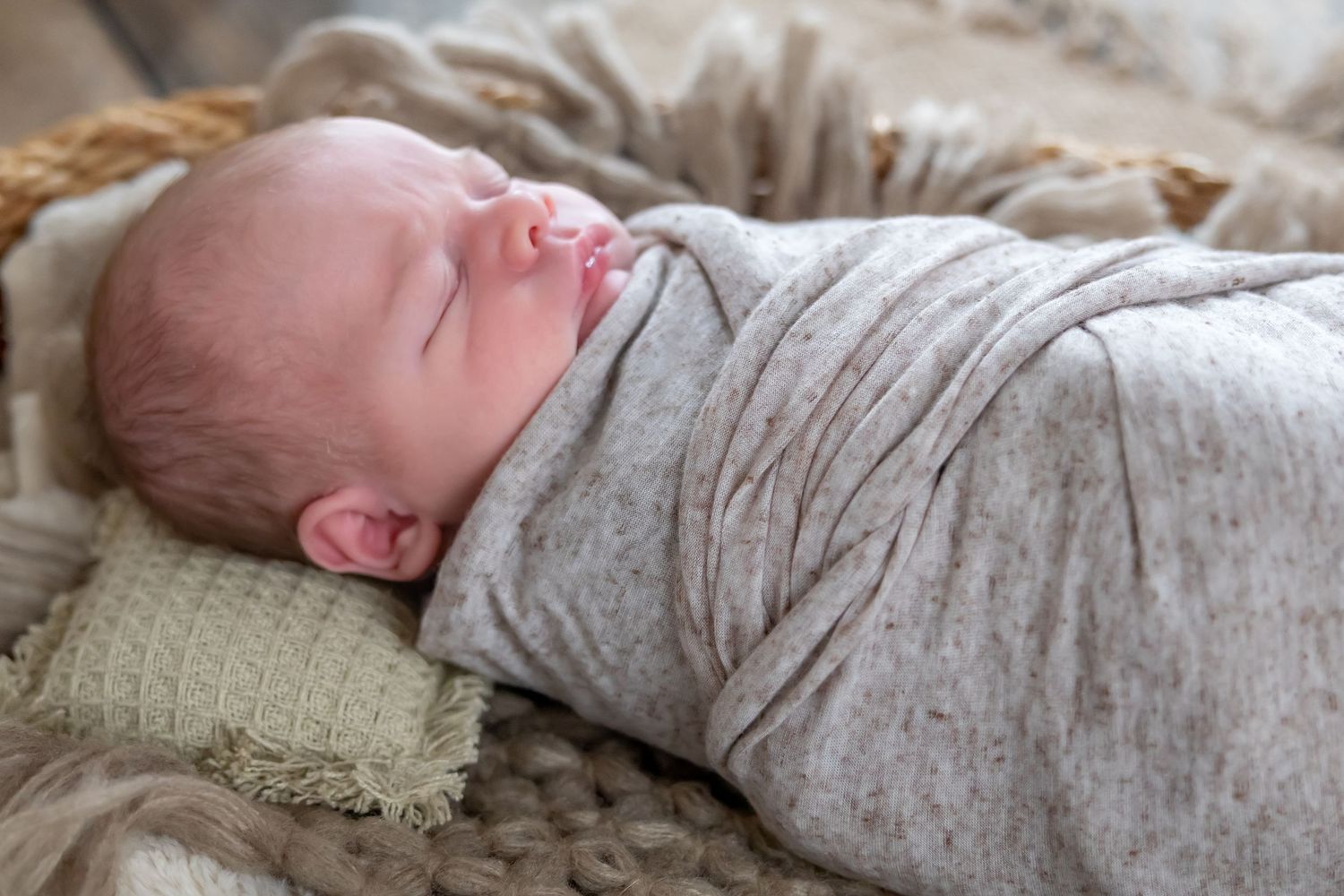 Sleeping newborn wrapped in soft gray swaddle rests on textured neutral bedding.