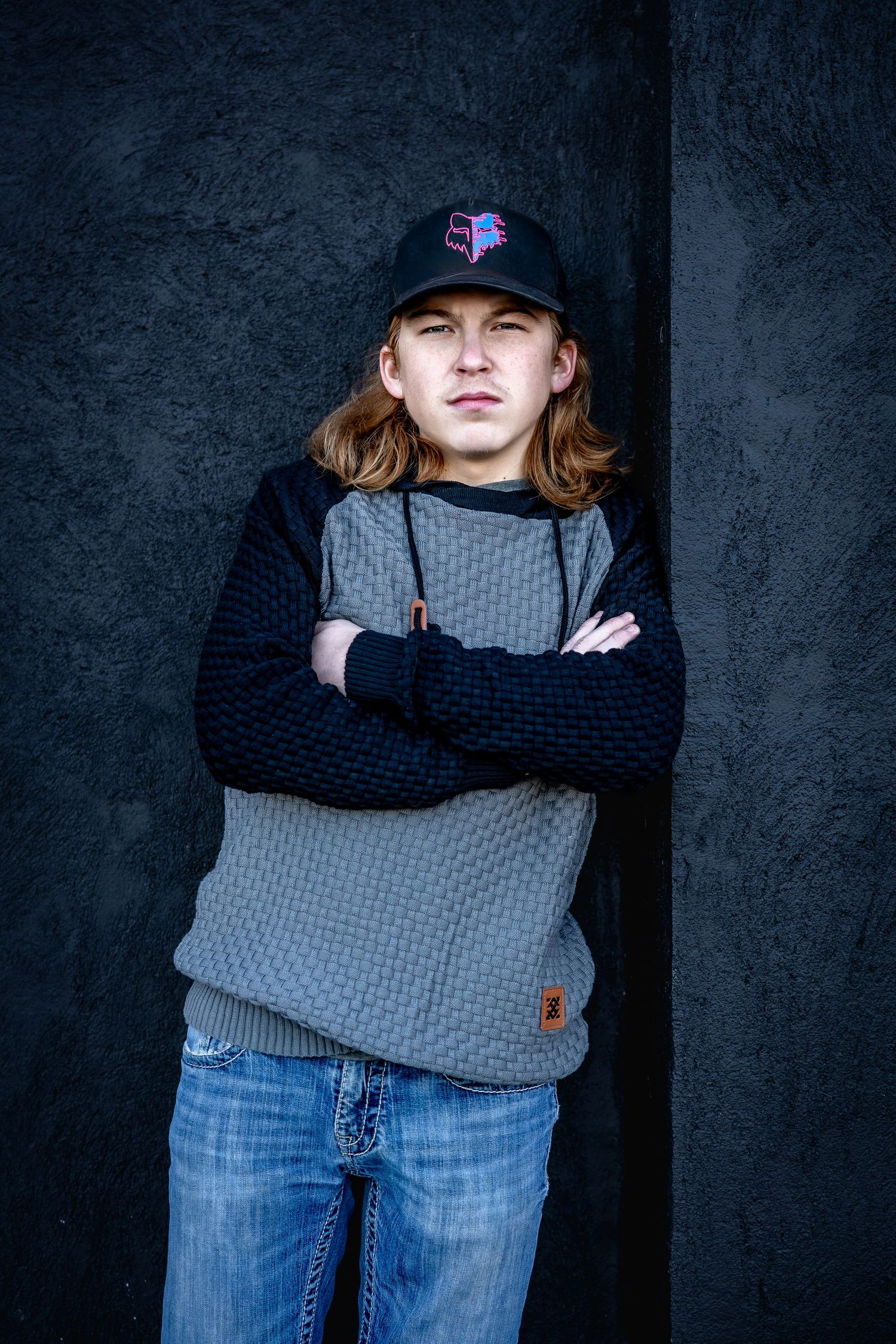 Studio portraits against dark background wearing grey sweater with navy sleeves and baseball cap.