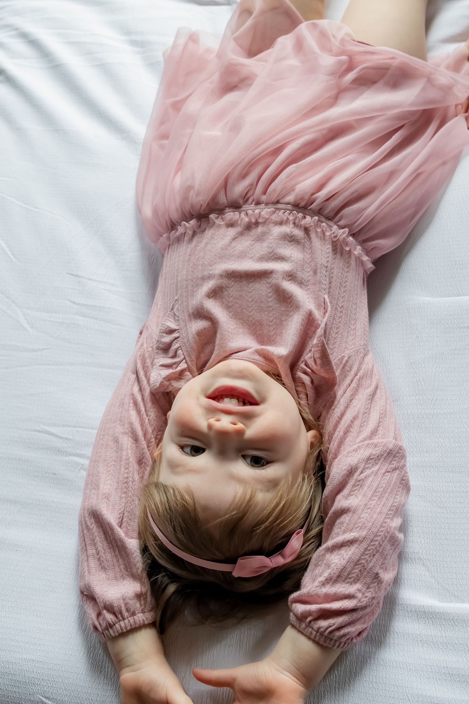 Baby in pink clothing lying on white sheets making playful expressions.