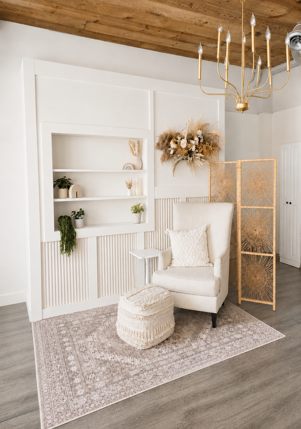 Boho chic living room with white wingback chair, woven pouf, built-in shelves, gold chandelier, and neutral pampas grass decor.