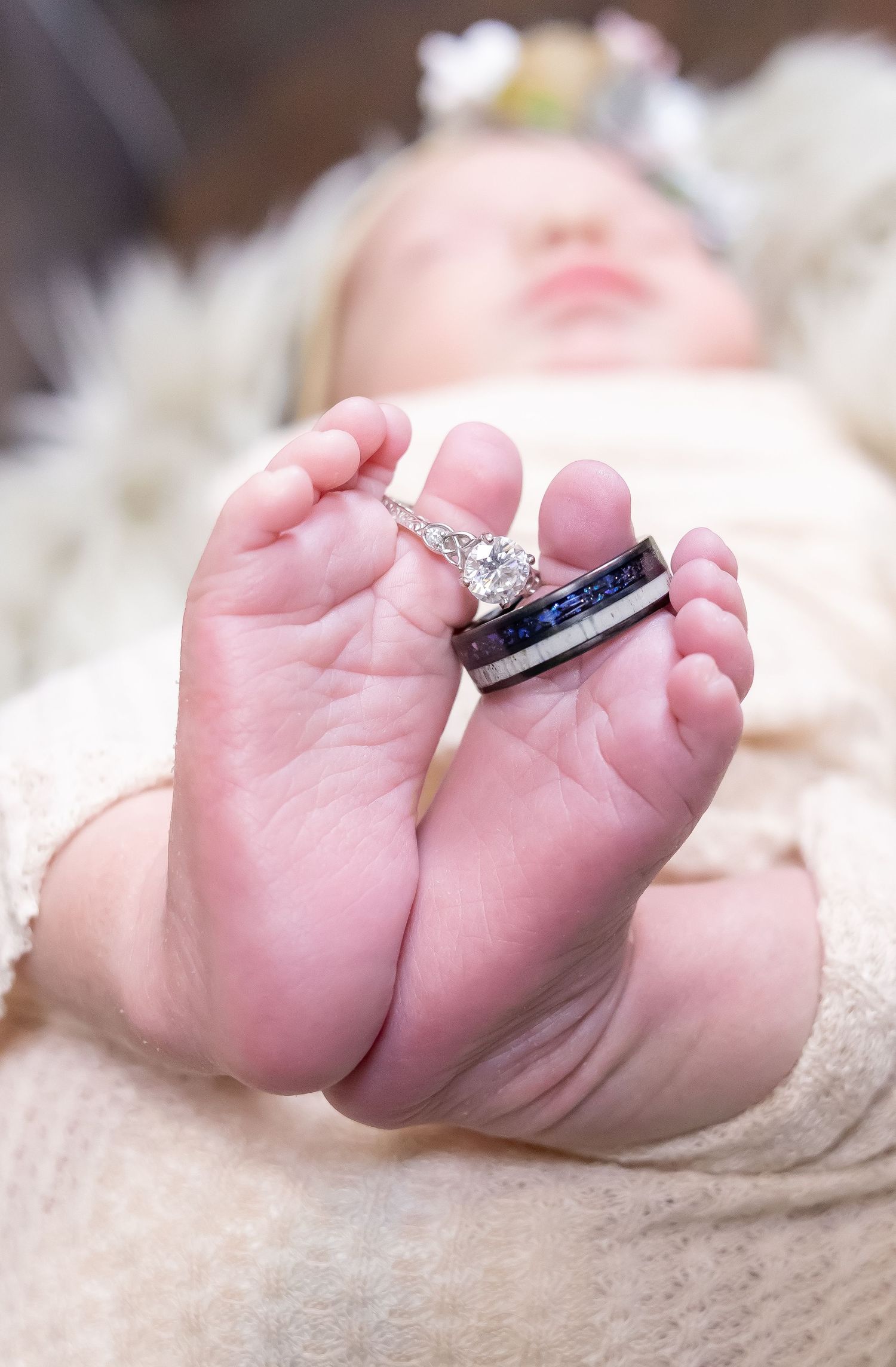 Tiny baby feet holding wedding rings against a white blanket in an artistic newborn photography pose.