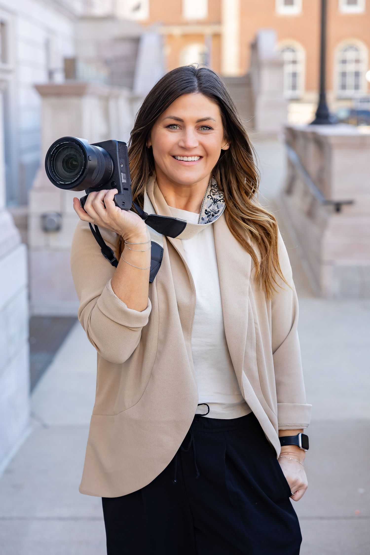 Professional photographer in beige blazer demonstrating camera poses against urban backdrop.