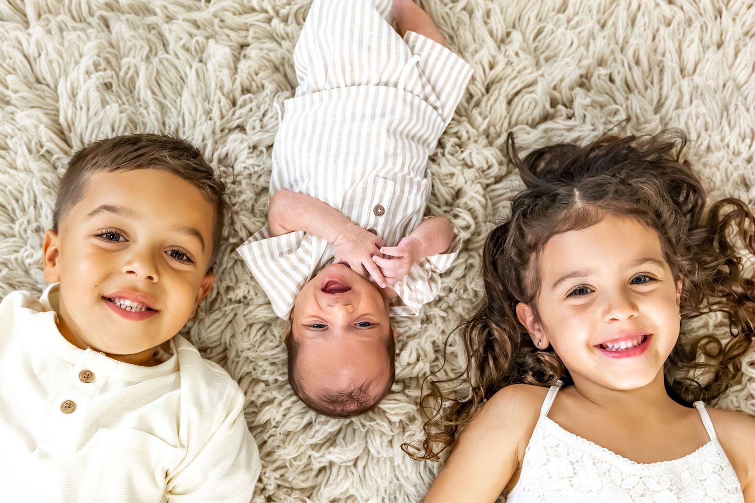 Family photo session on cream shag rug showing siblings with newborn.