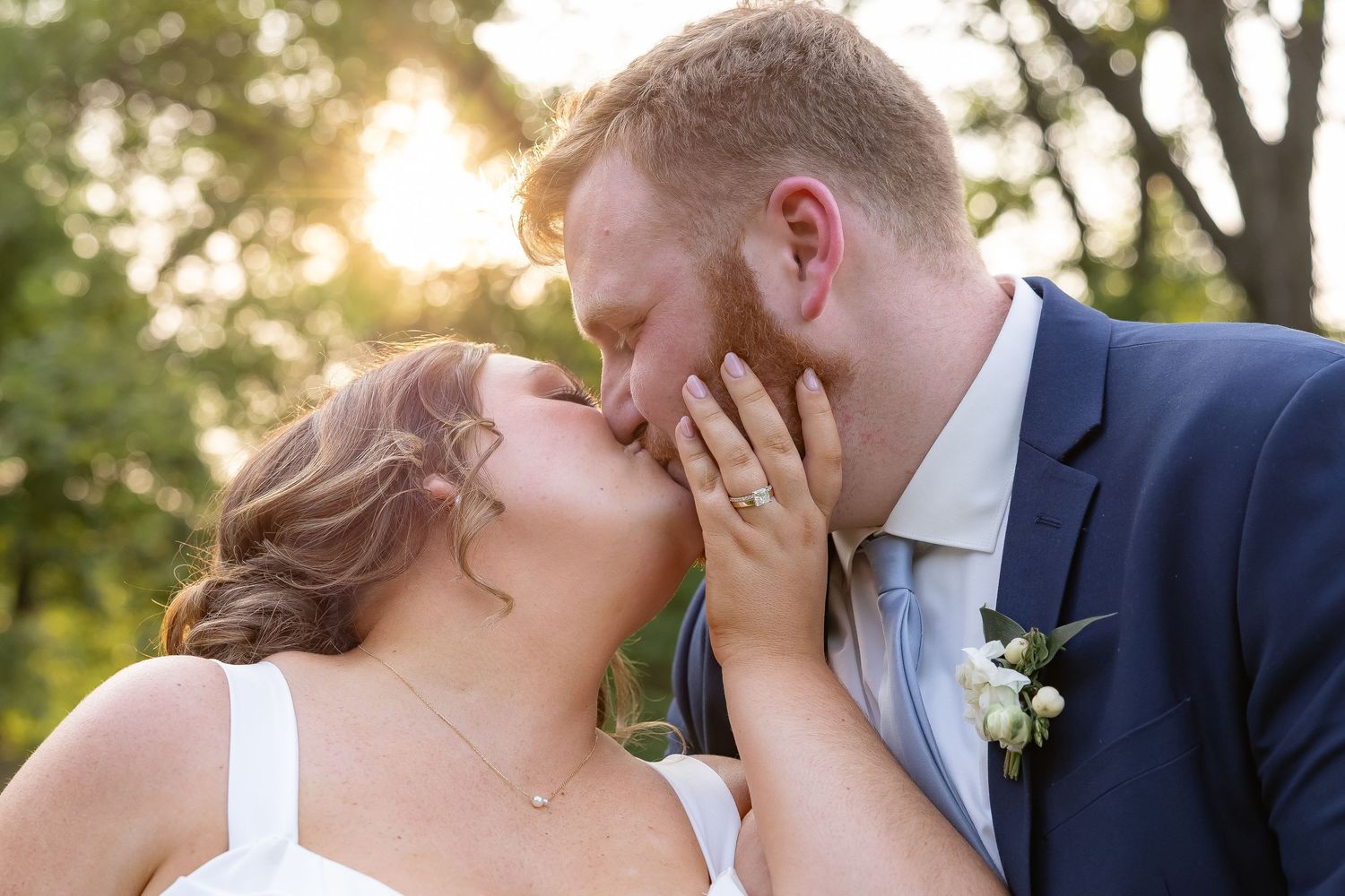 A romantic wedding couple shares an intimate kiss outdoors during golden hour sunlight in a garden setting.