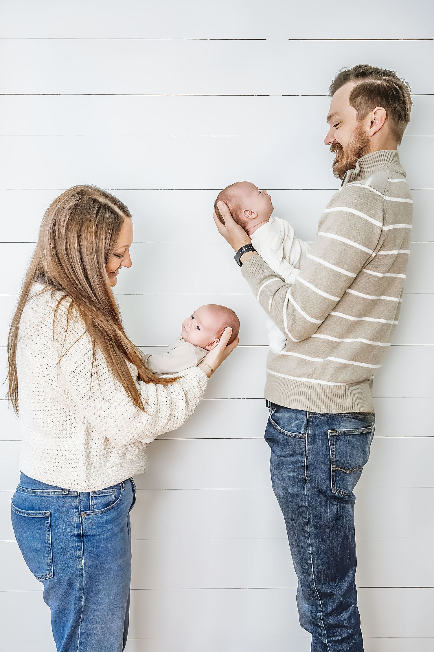 Parents in matching sweaters hold their twin babies while standing against a white shiplap wall.
