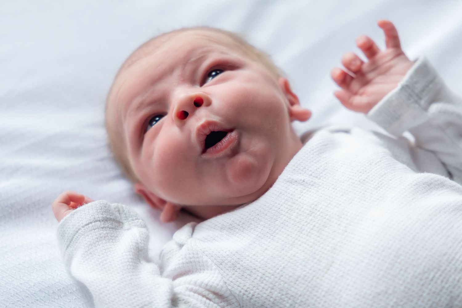Newborn baby in white outfit expressing joy on white bedding.