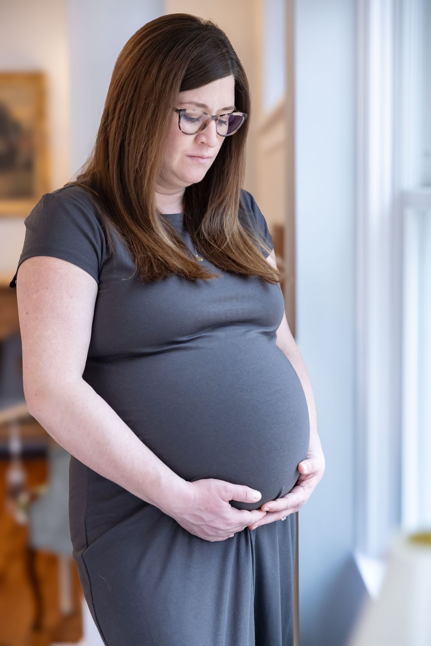 Maternity portrait of expectant mother in gray dress by window with natural lighting.