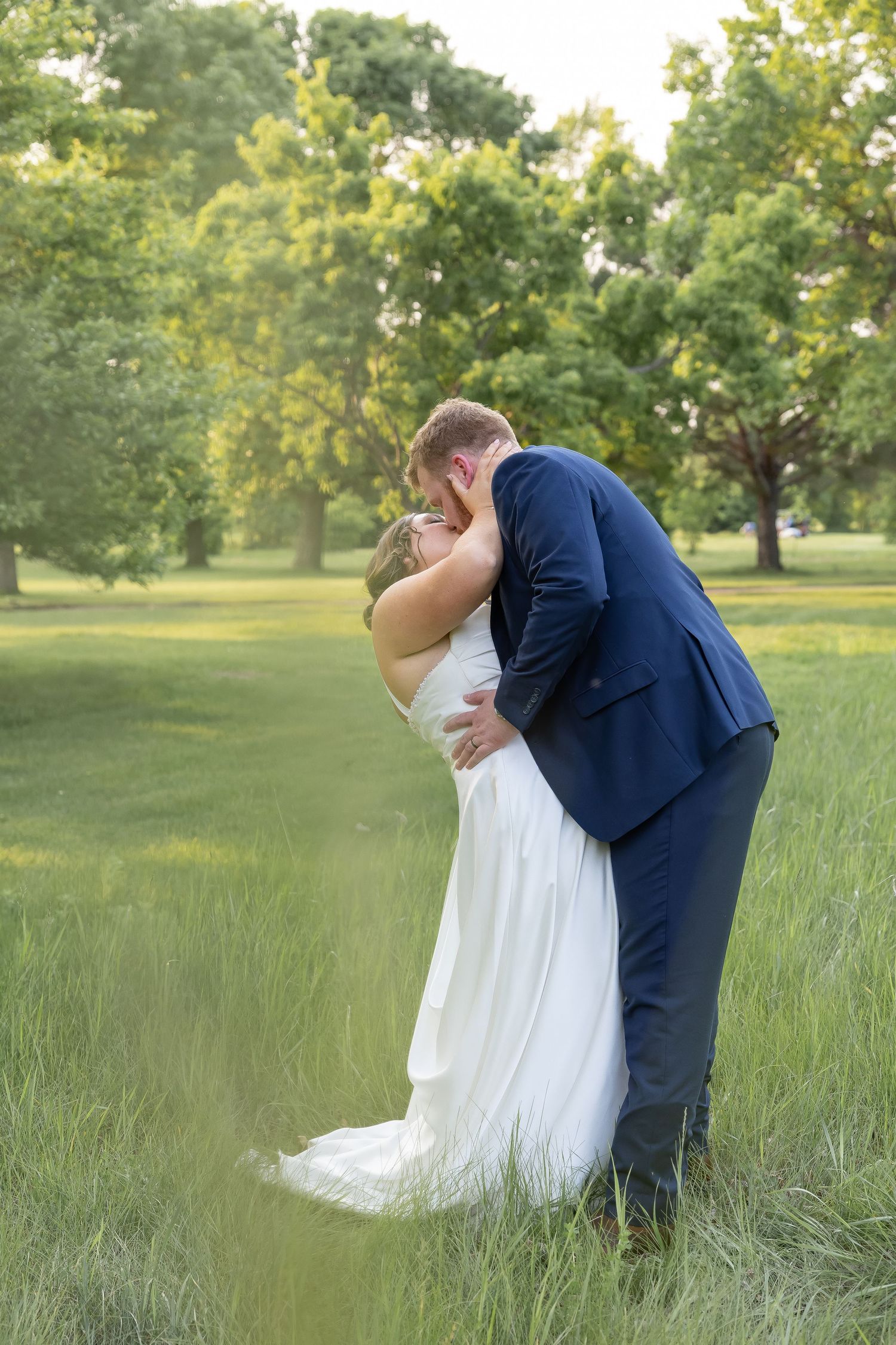 Romantic wedding couple sharing intimate kisses in a sunlit meadow with tall grass and trees in the background.