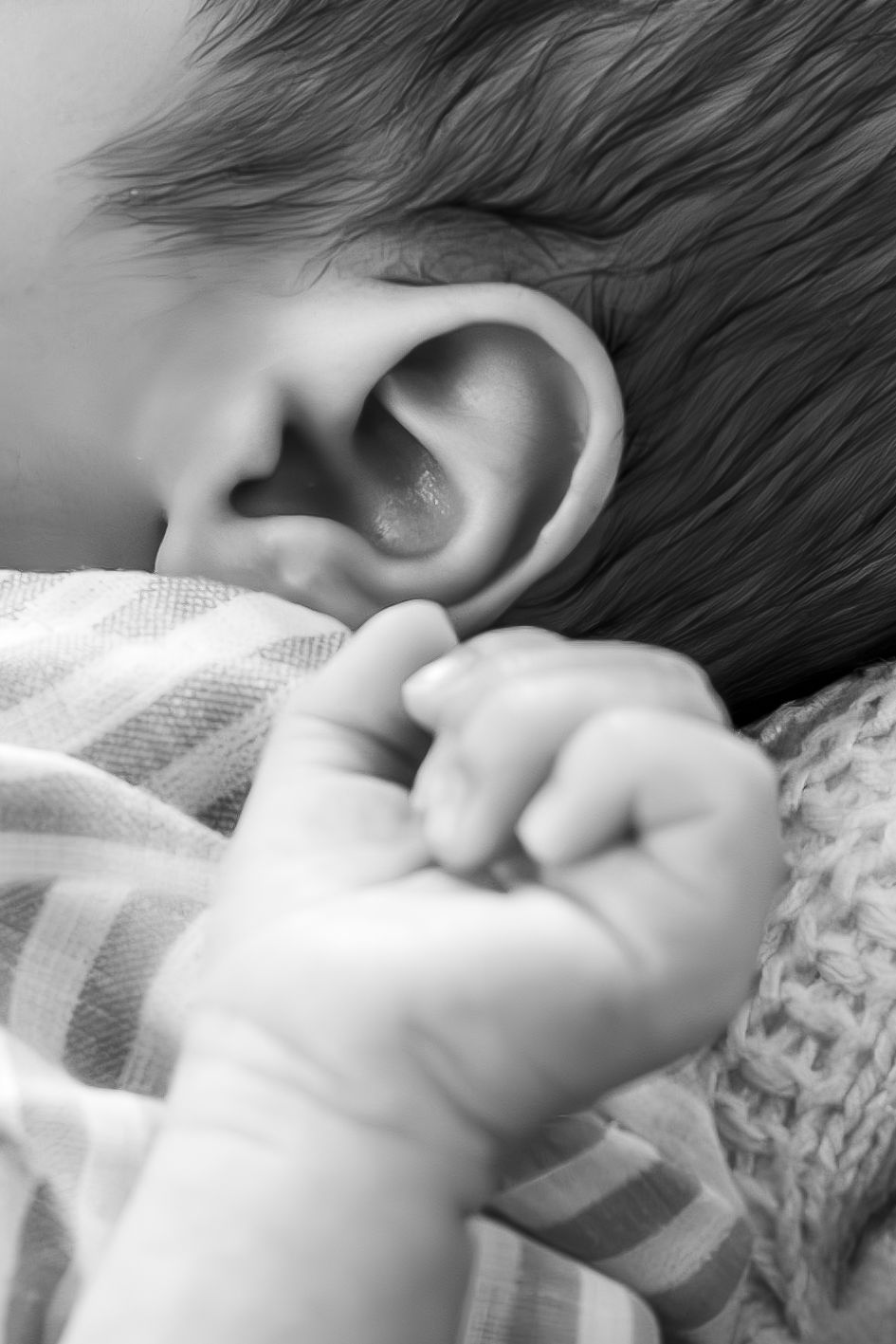 Black and white macro shot of baby's ear and tiny hand against striped fabric.