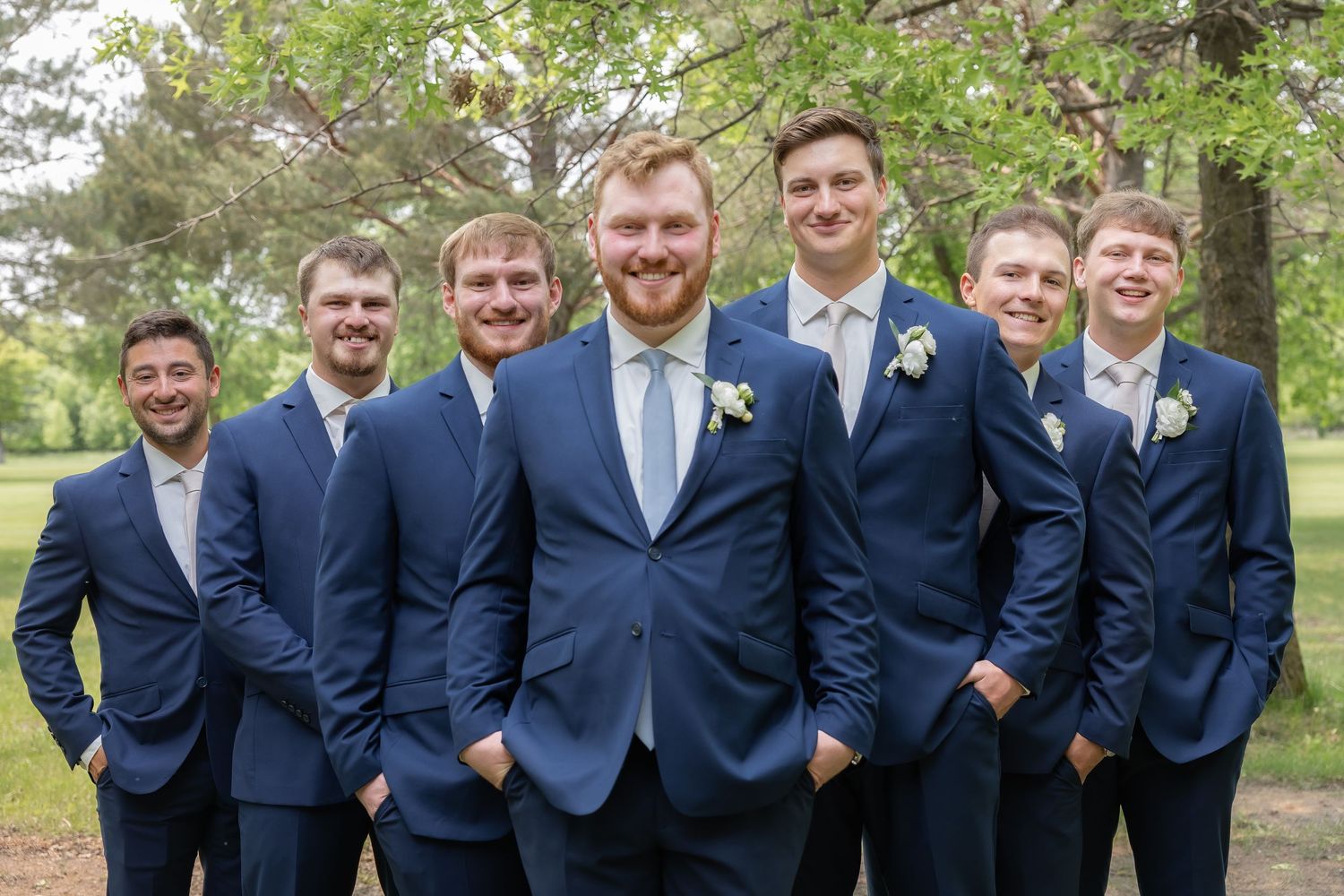 A group of groomsmen in navy blue suits stand together smiling outdoors in a park setting.