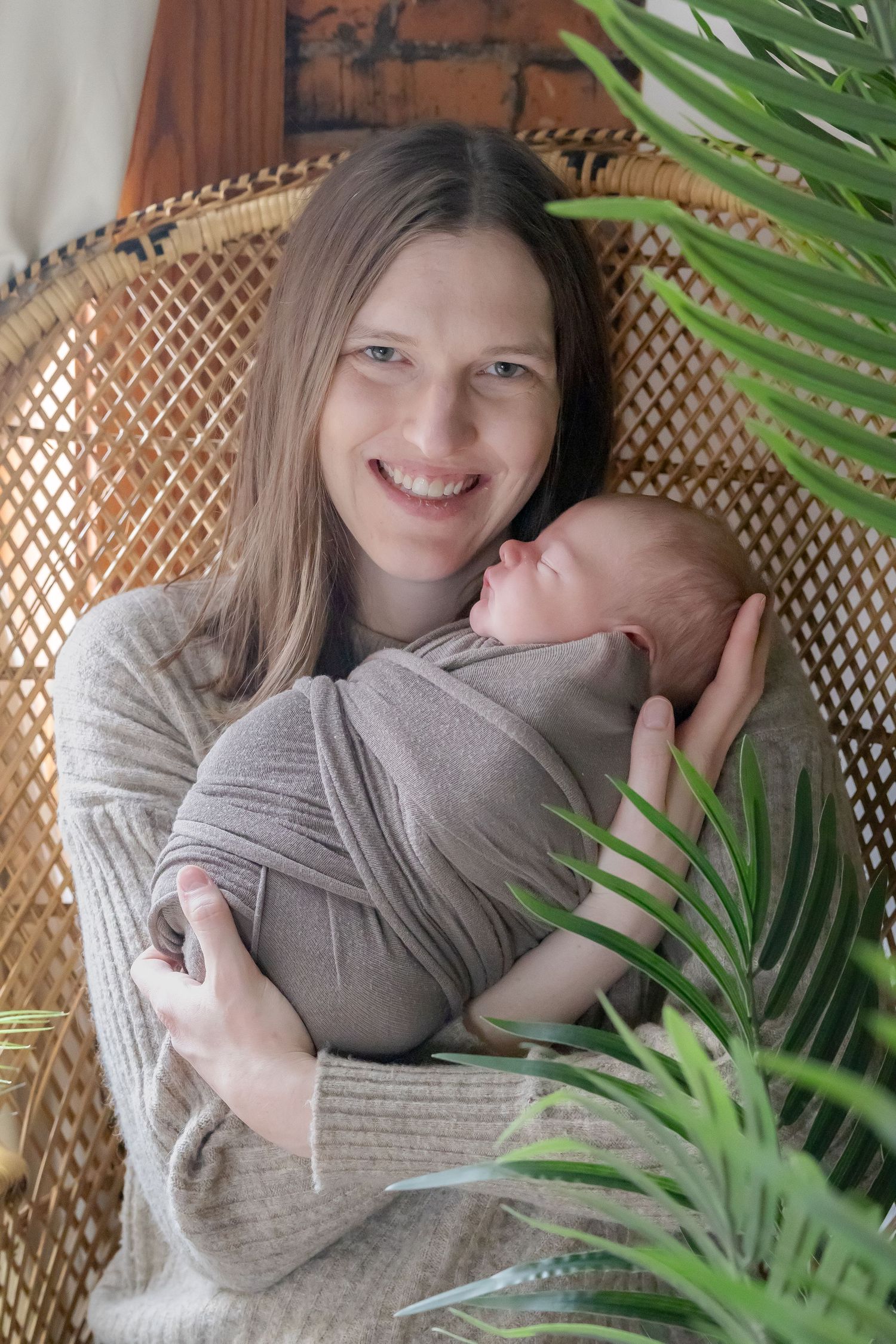 Parent and baby in gray wrap seated in rattan chair with tropical plants.