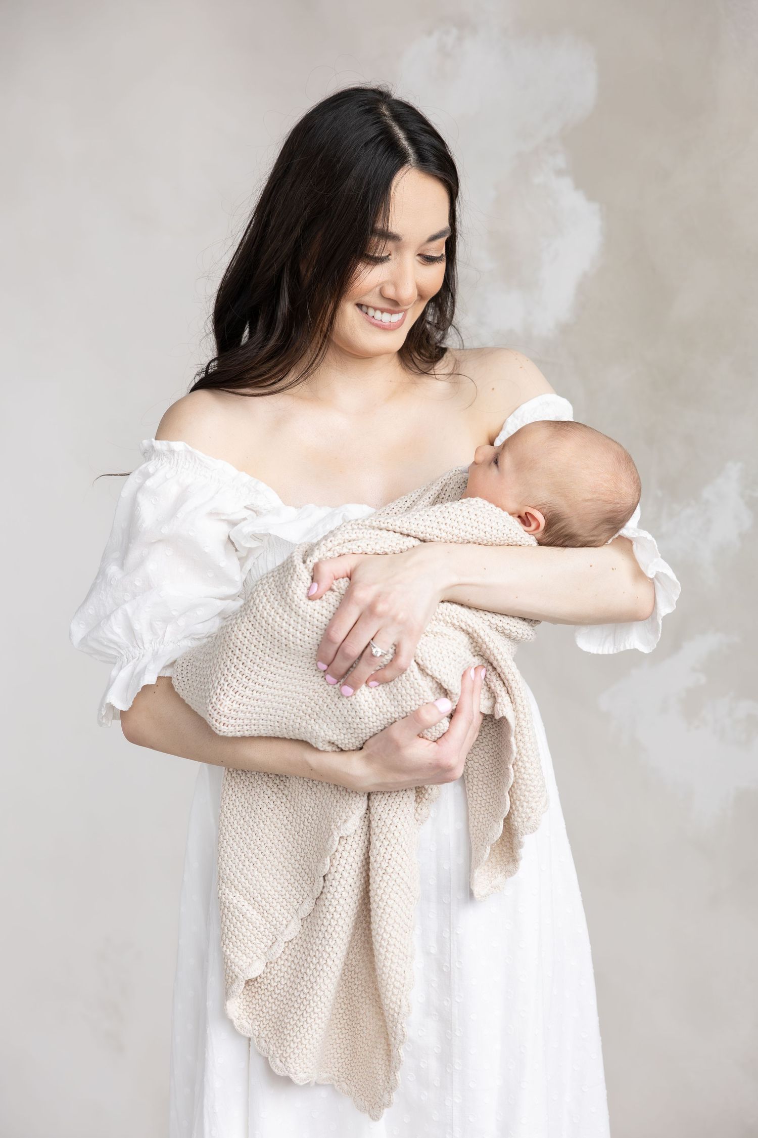 Smiling young mother in white dress tenderly holding sleeping newborn baby wrapped in beige knit blanket.