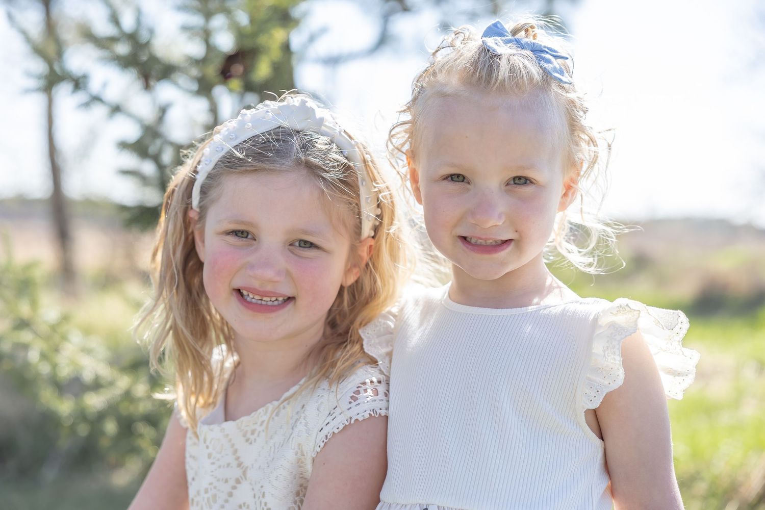 Children in white dresses pose together in sunny outdoor setting with pine trees.