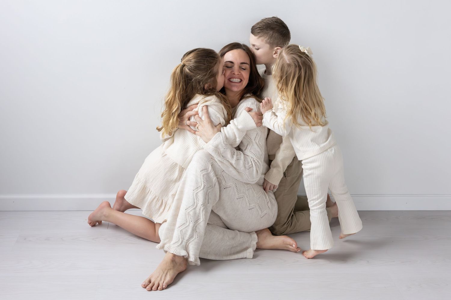 Family wearing cozy cream colored outfits embrace while sitting on floor against white wall.