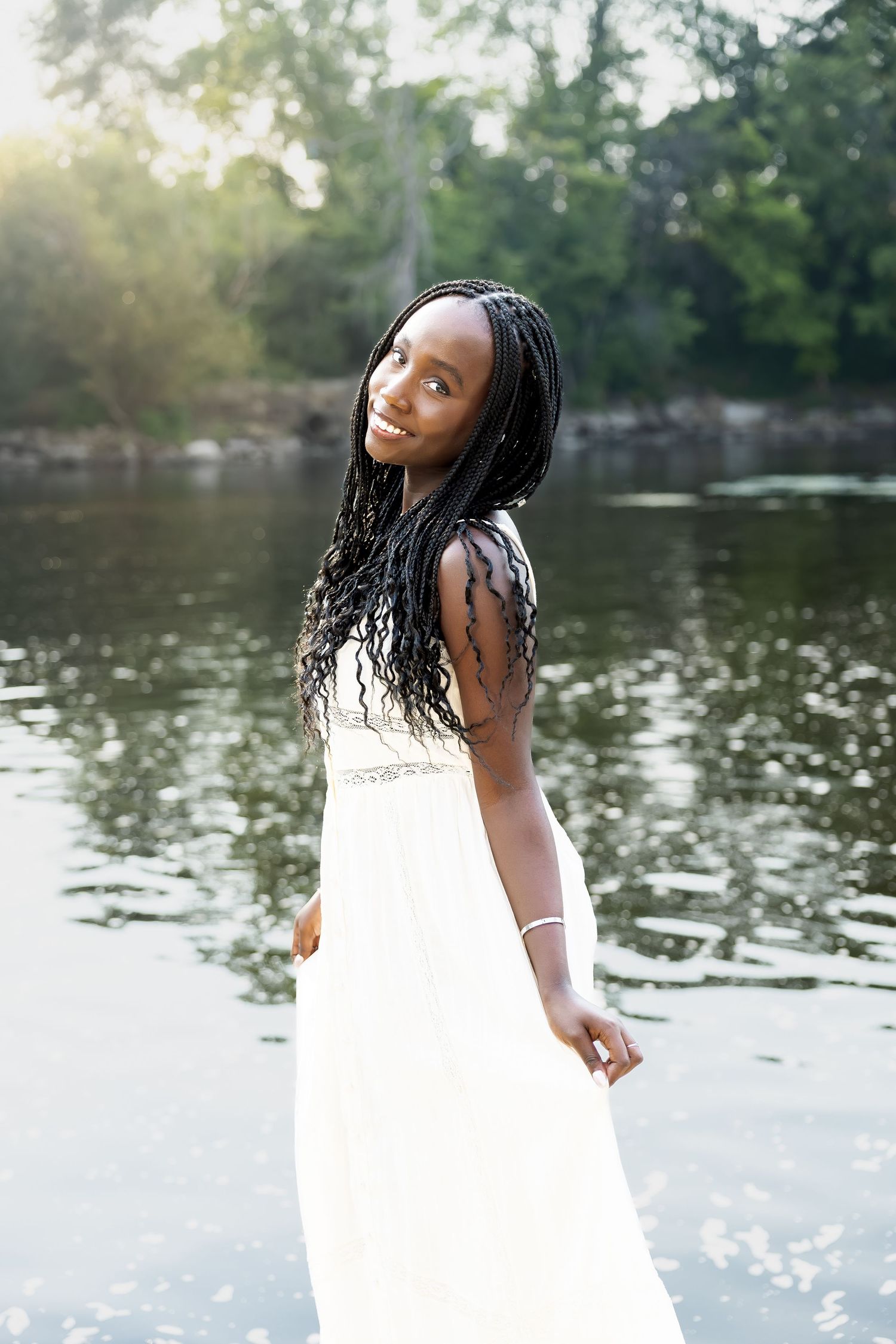 A person in a white dress poses gracefully by a serene lake with sunlight reflecting on the water.