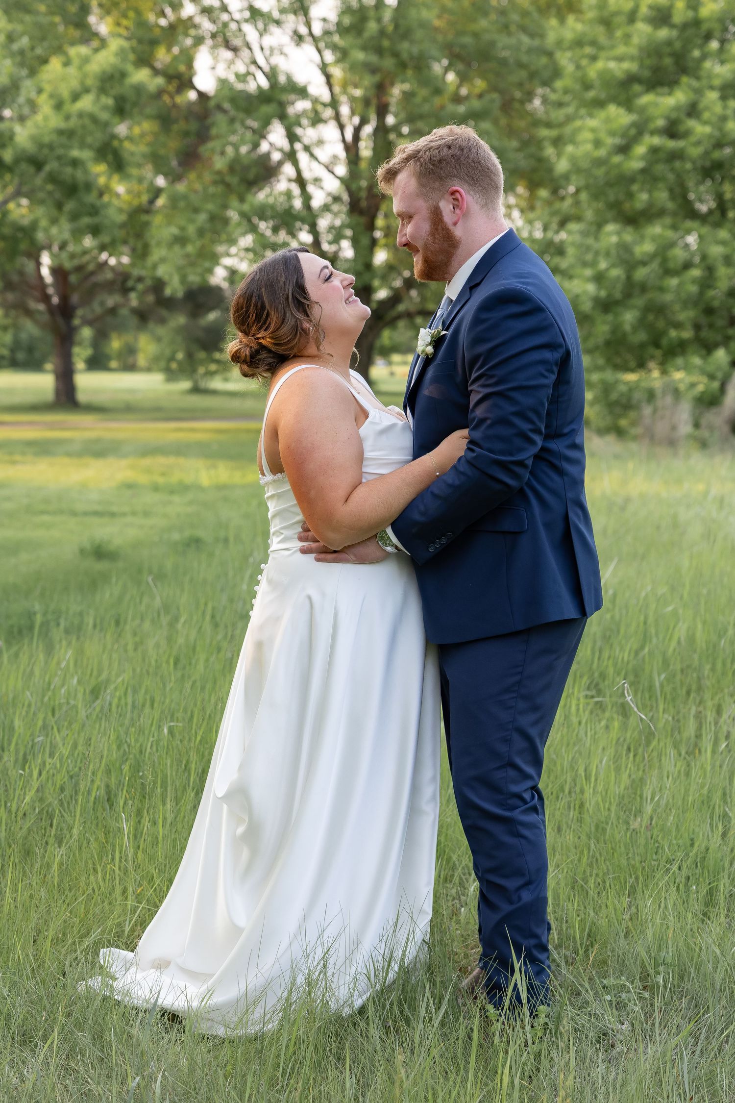 A romantic wedding couple shares an intimate moment together in a sunlit meadow with tall grass and trees.