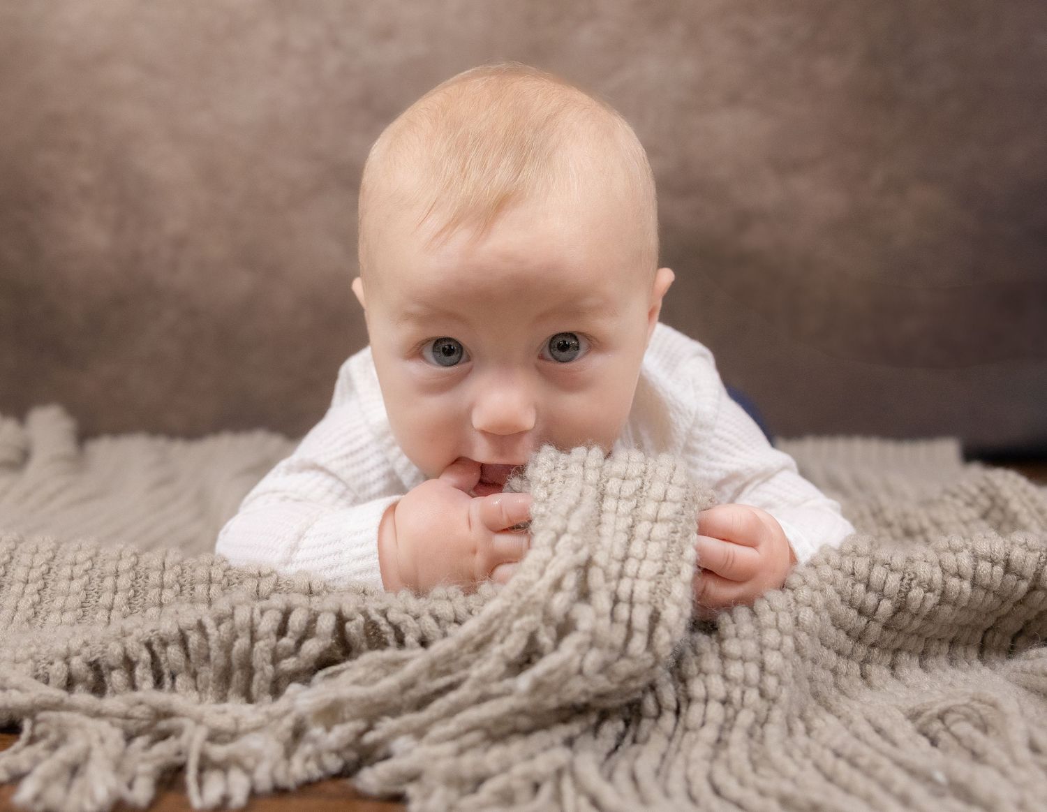 Baby in white outfit lying on gray knit blanket during professional photo session.