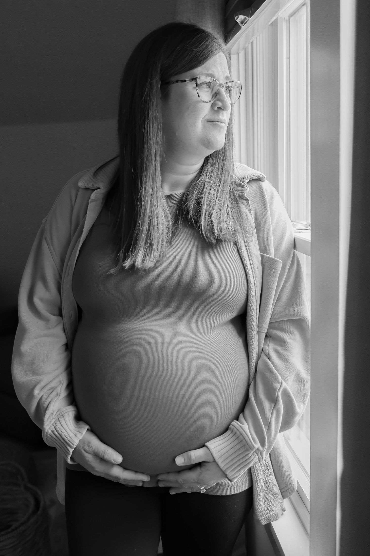 Black and white maternity portrait of expectant mother gazing out window.