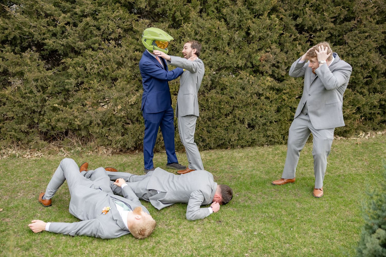 Groomsmen in light gray suits playfully pose and pretend to fight while one lies on grass during wedding photoshoot.