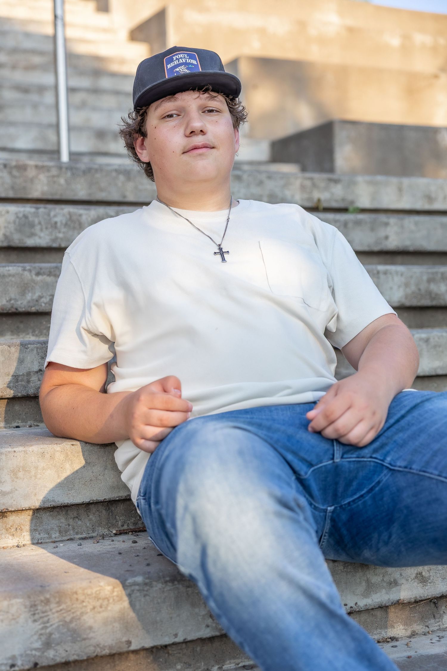 Casual portrait on concrete steps wearing baseball cap, white shirt and cross necklace.