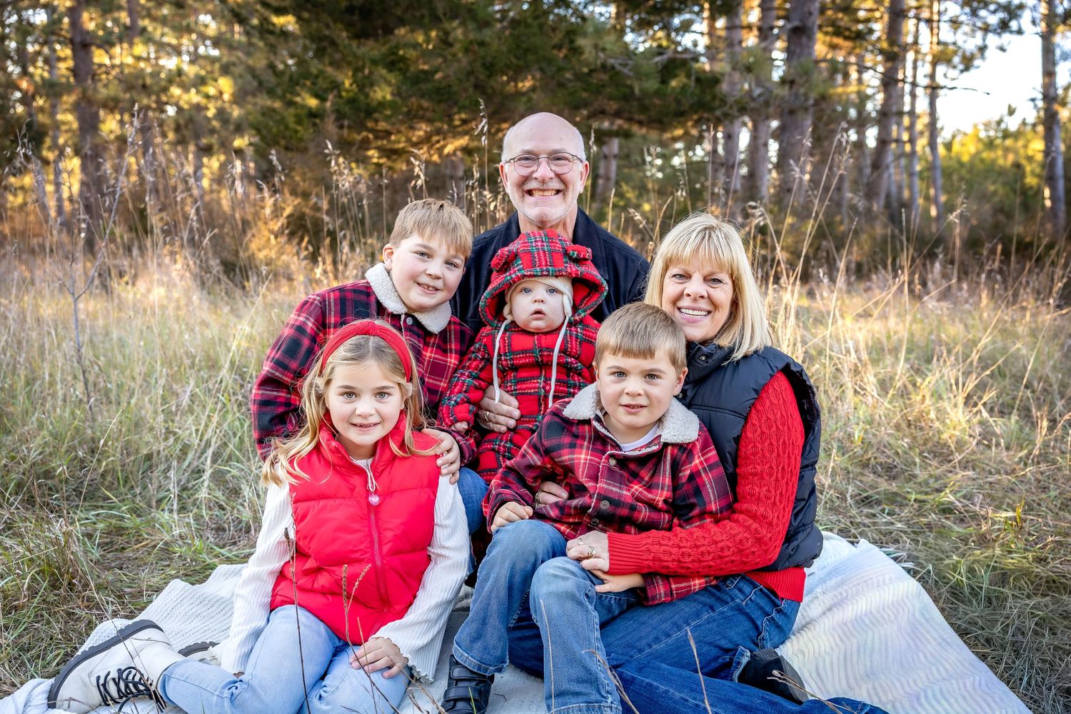 A family in coordinating red and plaid outfits poses together on a blanket in a pine forest.