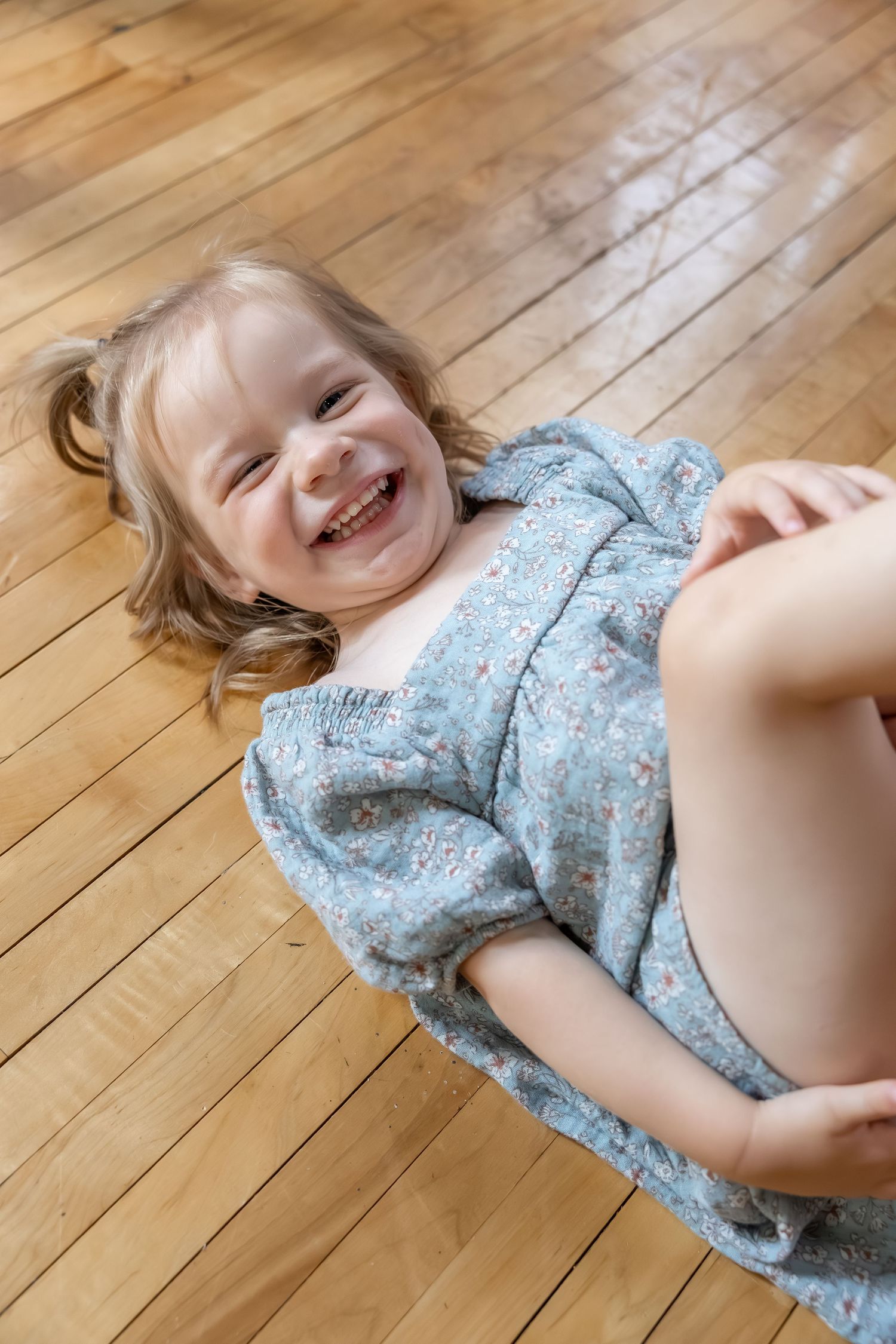 A child in a blue floral dress lies laughing on hardwood flooring.