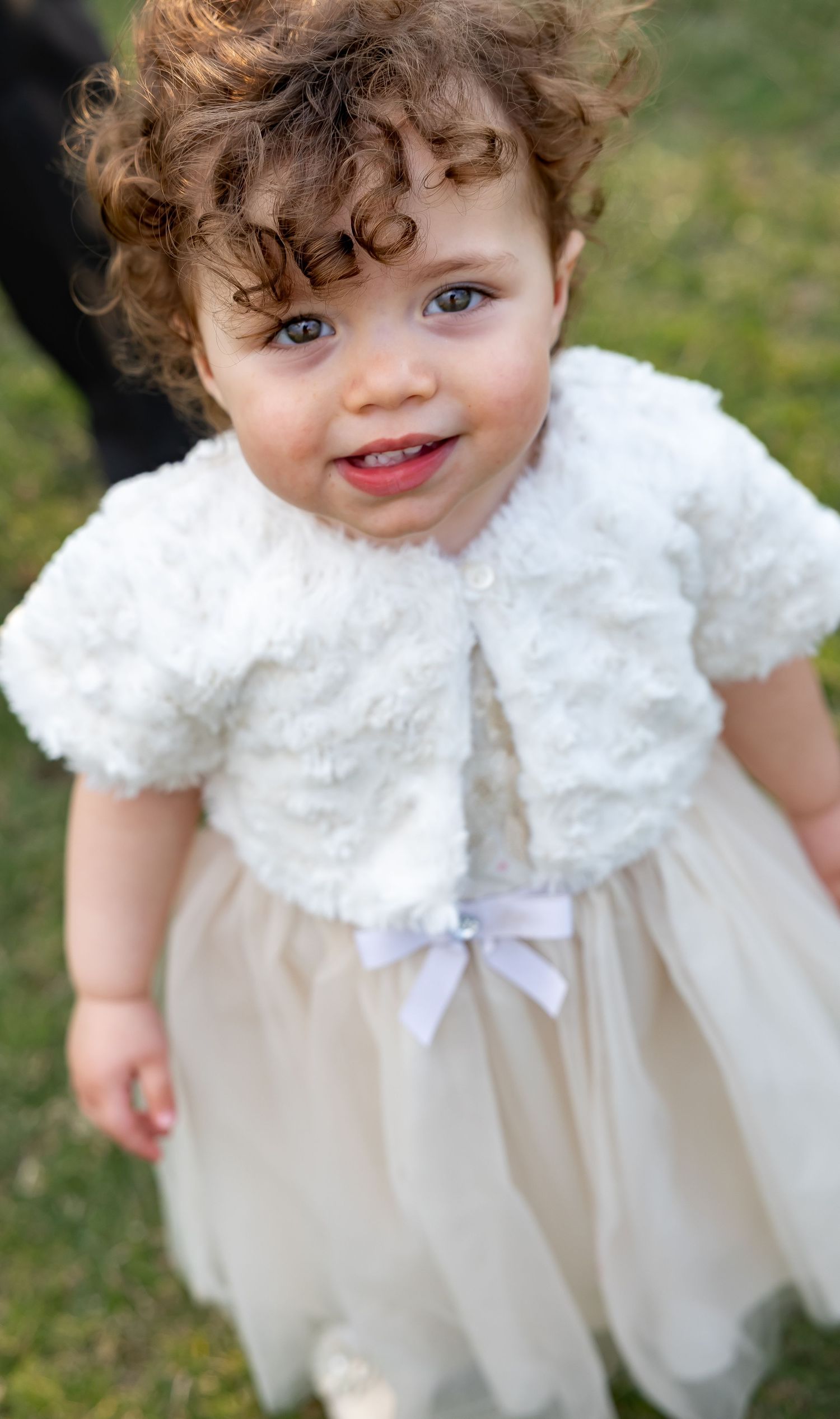 A young child in a white lace dress with curly hair stands on grass.