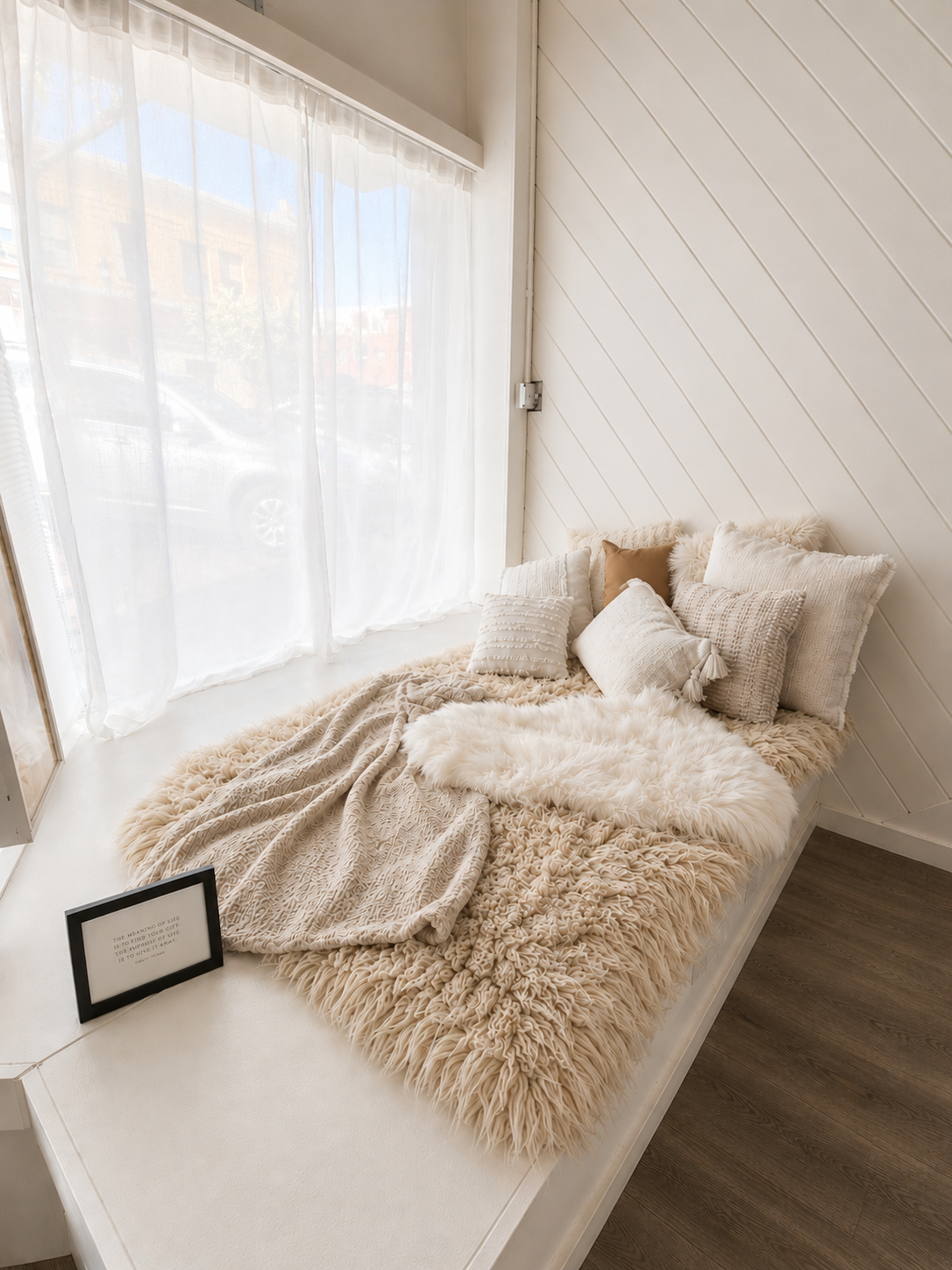 Cozy window seat with fluffy white pillows, beige throws, and sheer curtains in a bright minimalist room.