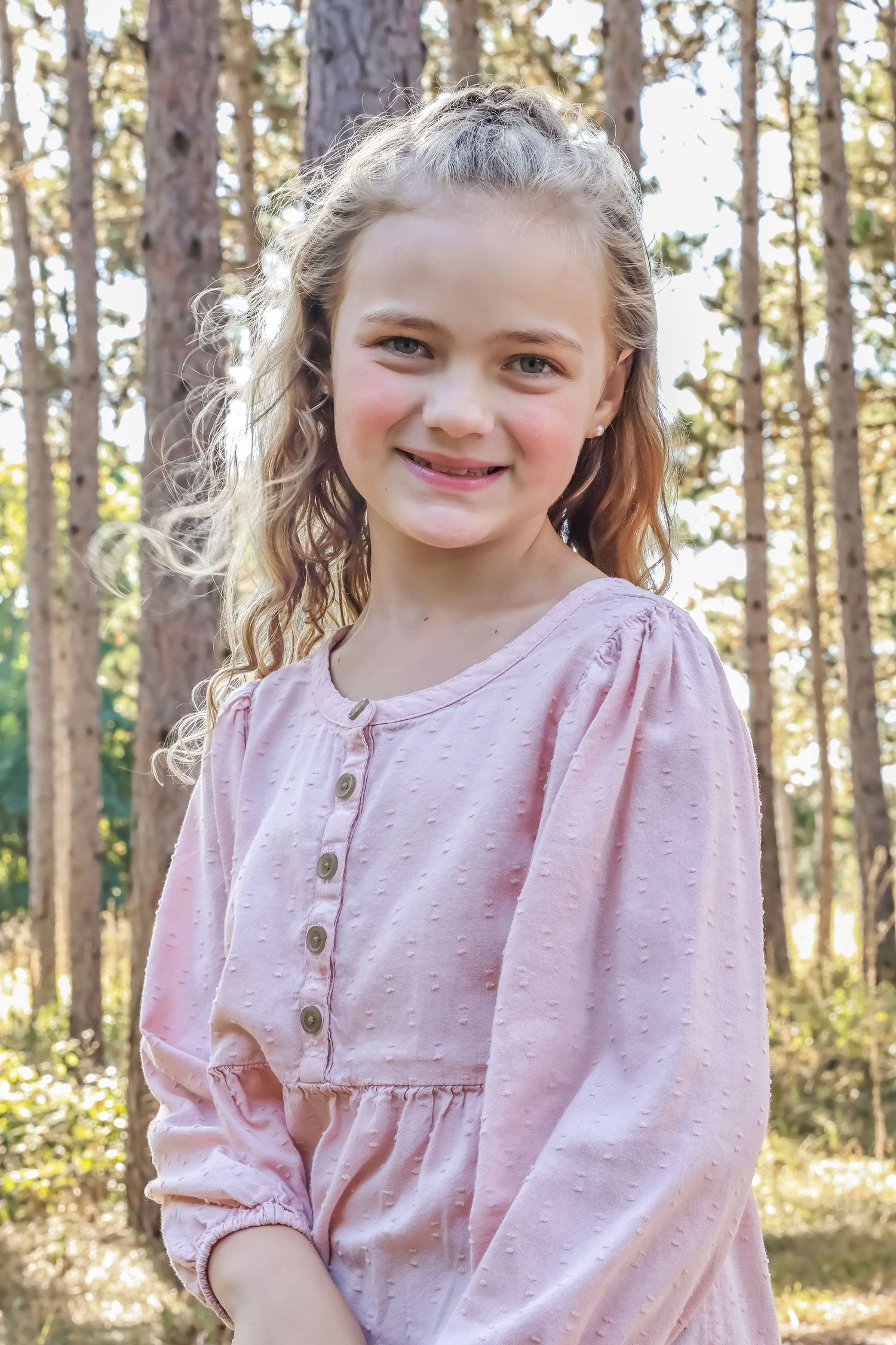 Young girl in a pink dress poses outdoors in a sunlit forest setting.