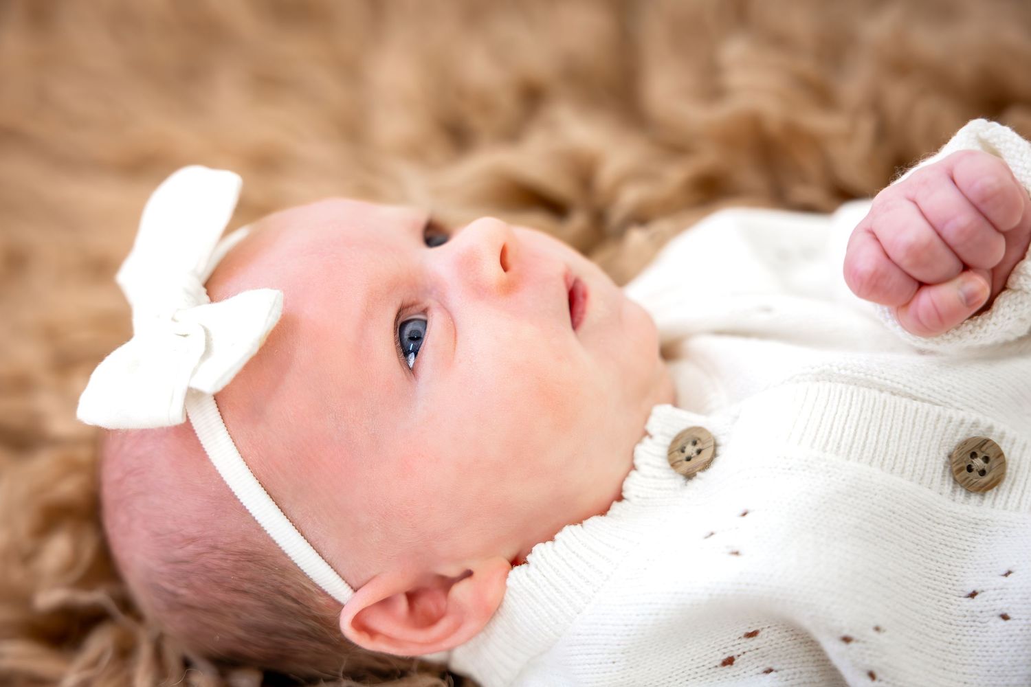 A baby in a white onesie lies on brown autumn leaves while smiling and playing outdoors.
