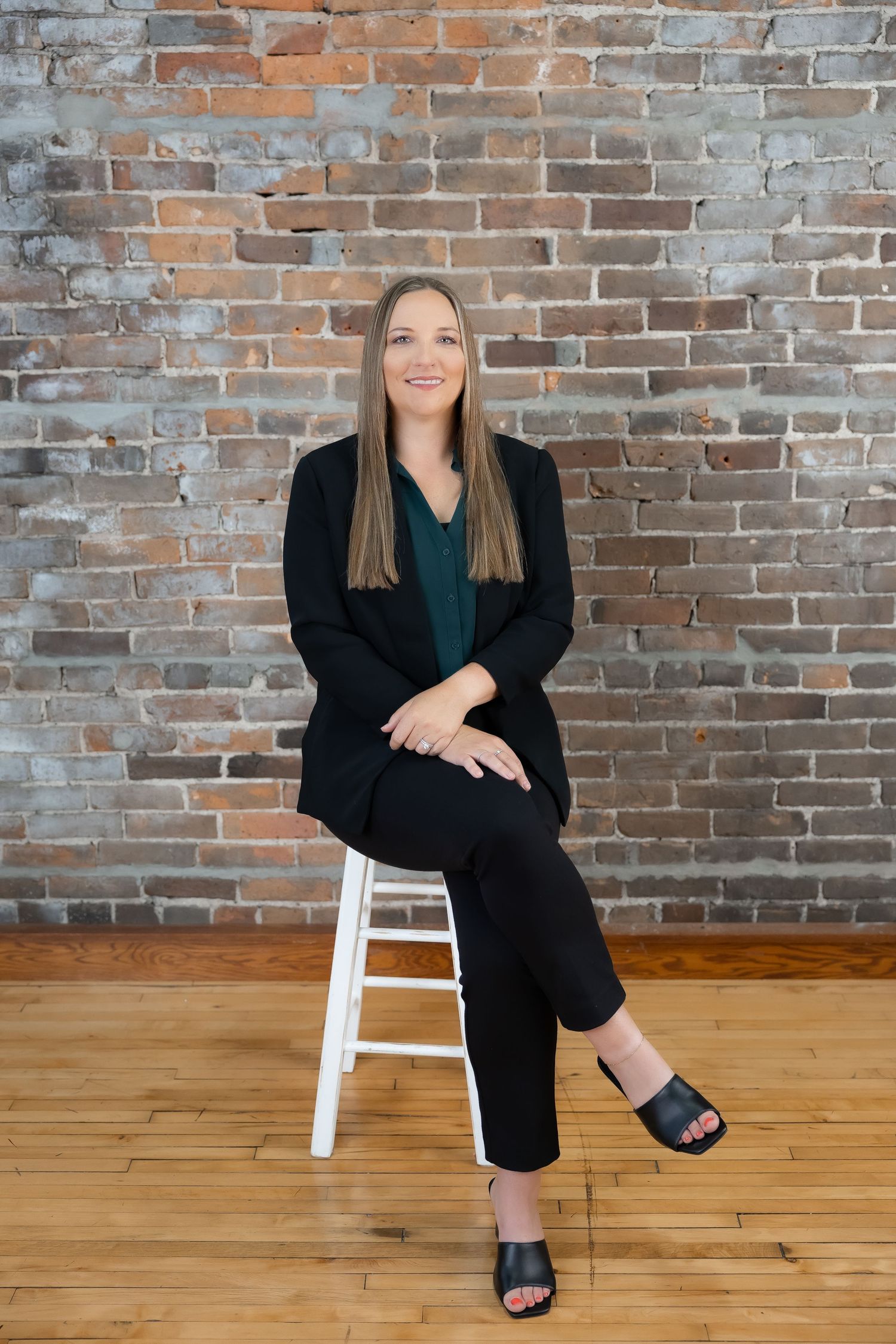 Professional portrait in business attire seated on white stool against exposed brick wall.