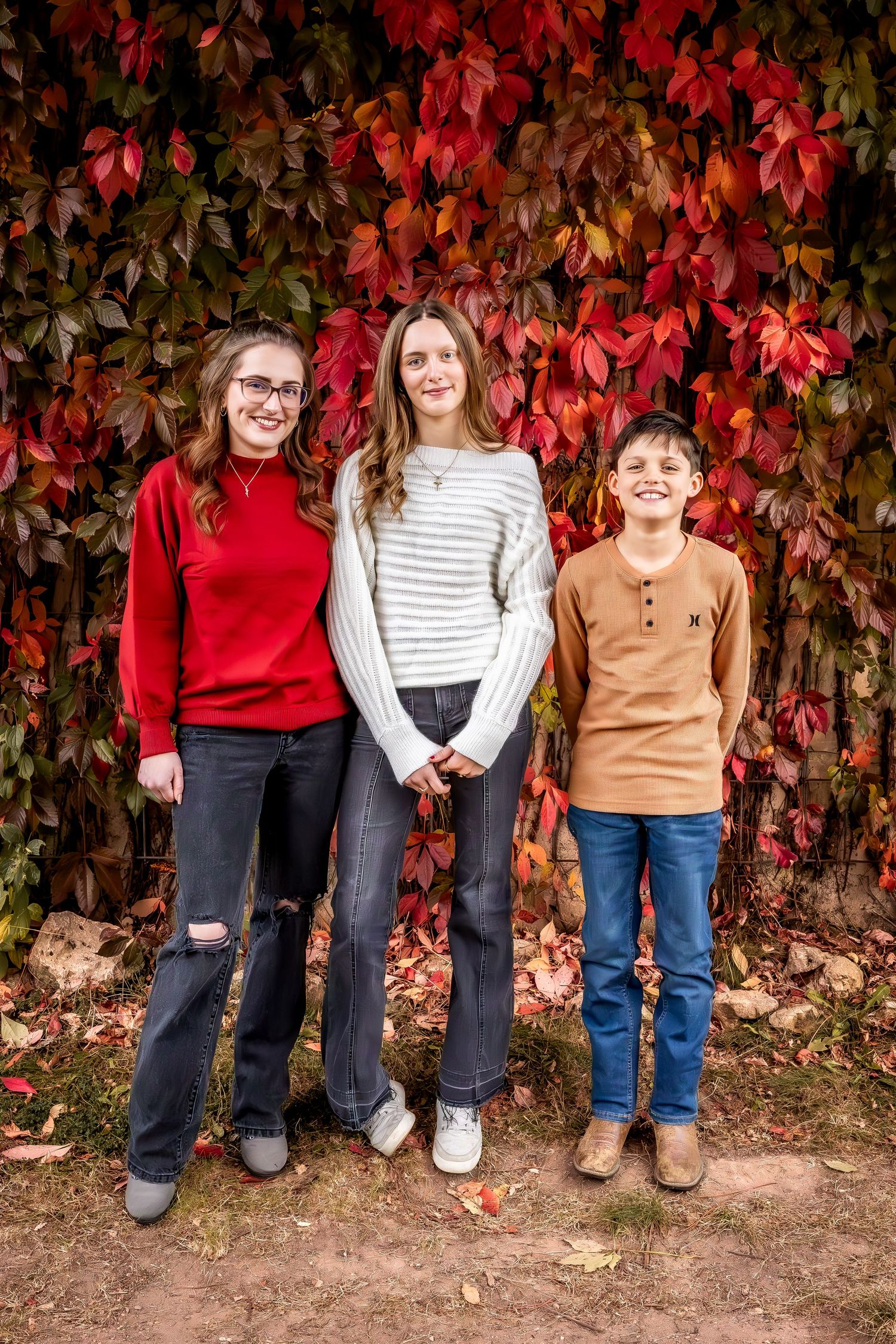 Three people pose against a wall of vibrant red fall leaves wearing coordinated casual outfits.