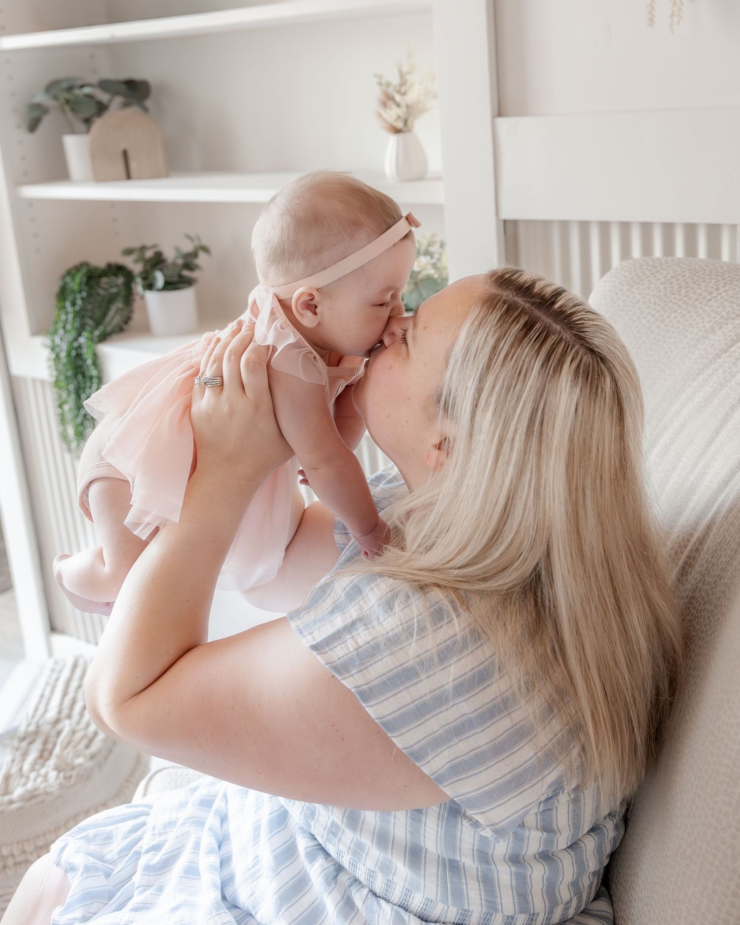 A blonde mother kisses her baby girl in a pink dress while sitting in a cozy nursery chair.