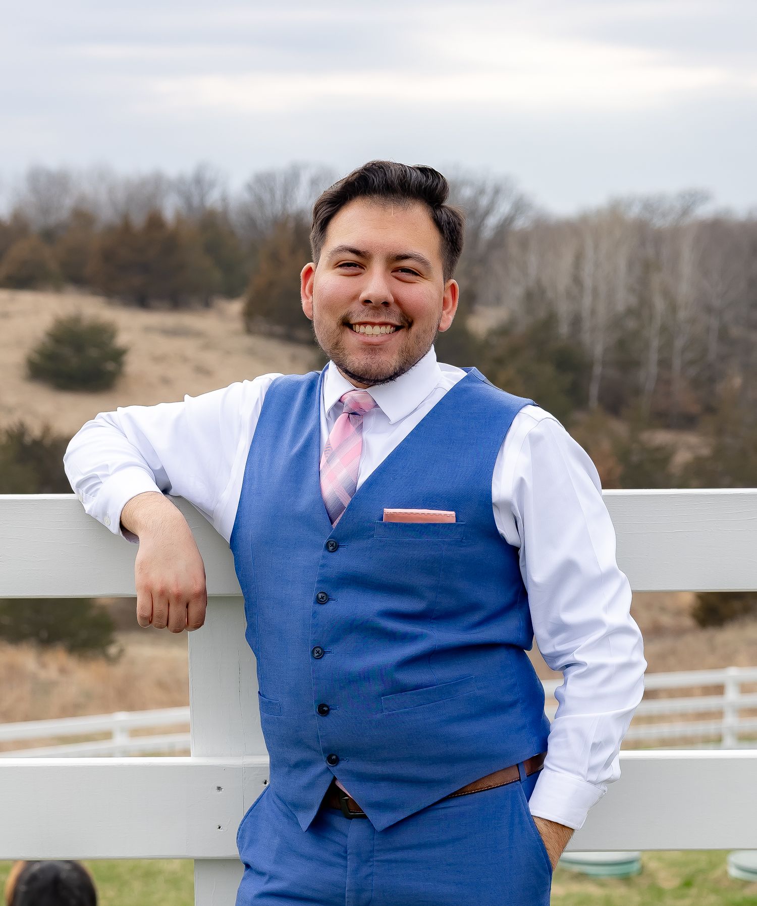Person wearing blue vest and white shirt poses casually by white fence in outdoor setting.
