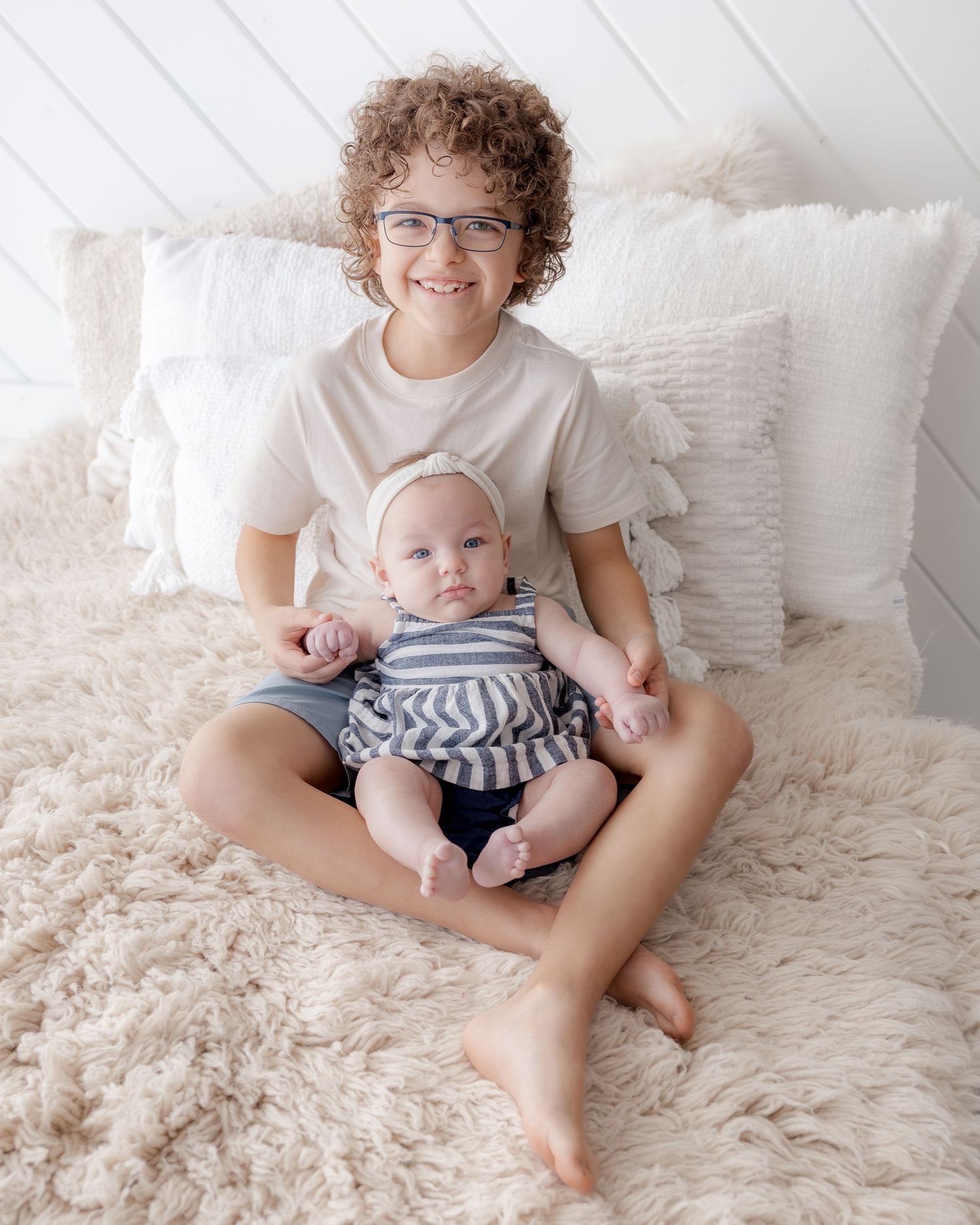 Smiling boy with curly hair and glasses holds a baby in striped outfit on a fluffy beige rug with white pillows.