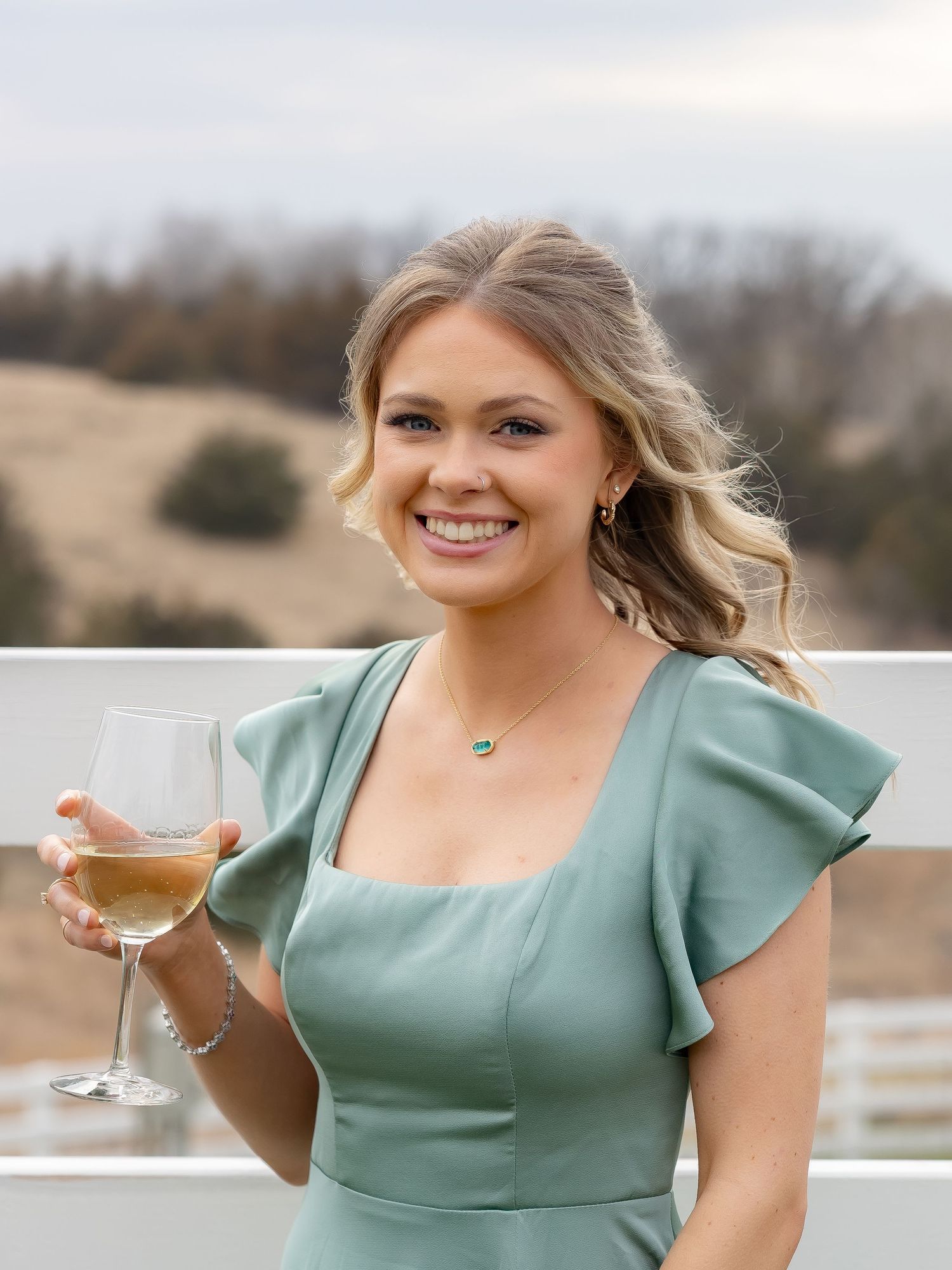 Person in mint dress holding wine glass while posing against rustic outdoor backdrop.