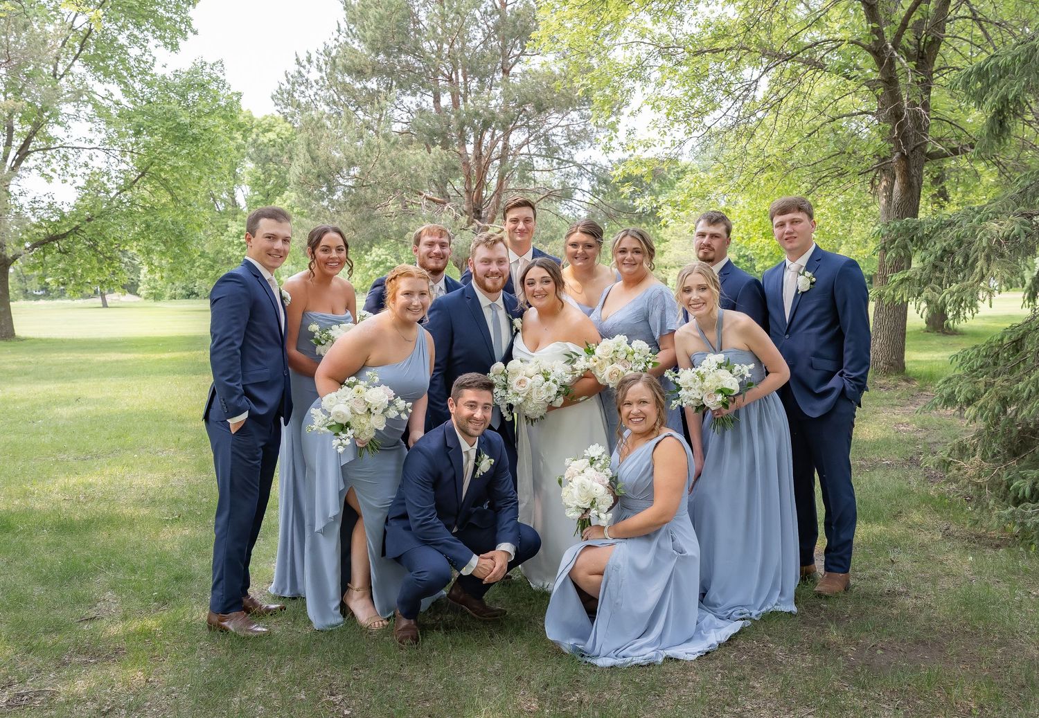 Wedding party in navy suits and light blue dresses poses together outdoors under trees in a natural setting.