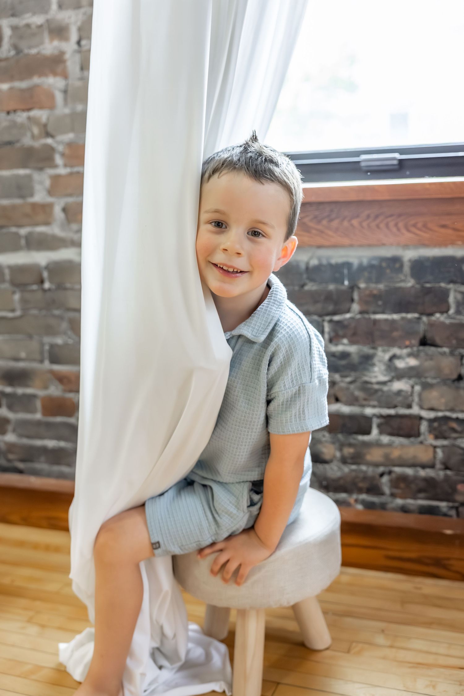 A child in striped clothing sits on a stool near exposed brick wall and white curtains.